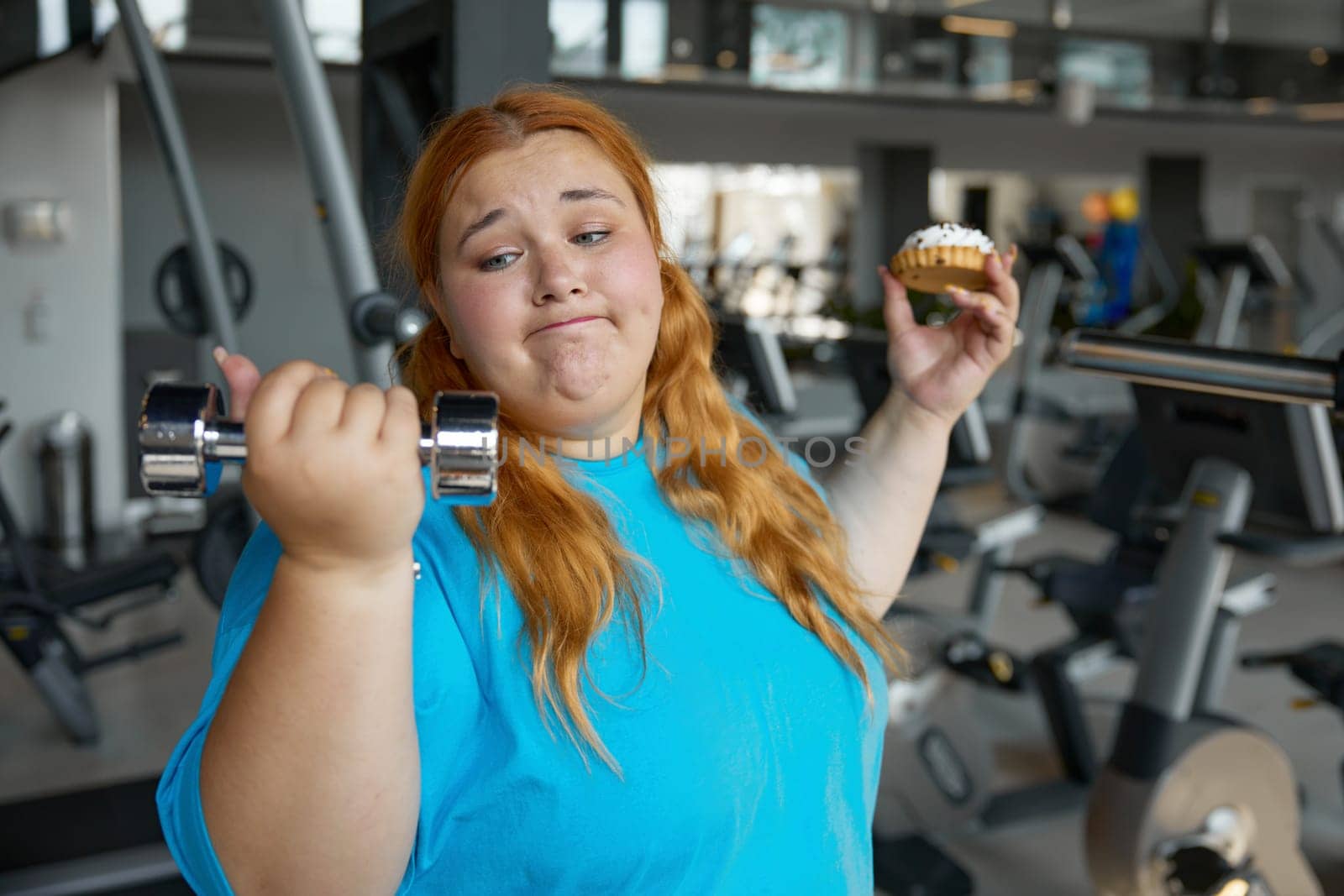 Portrait of comic overweight woman holding dumbbell and donut. Sport and fitness workout for slimming