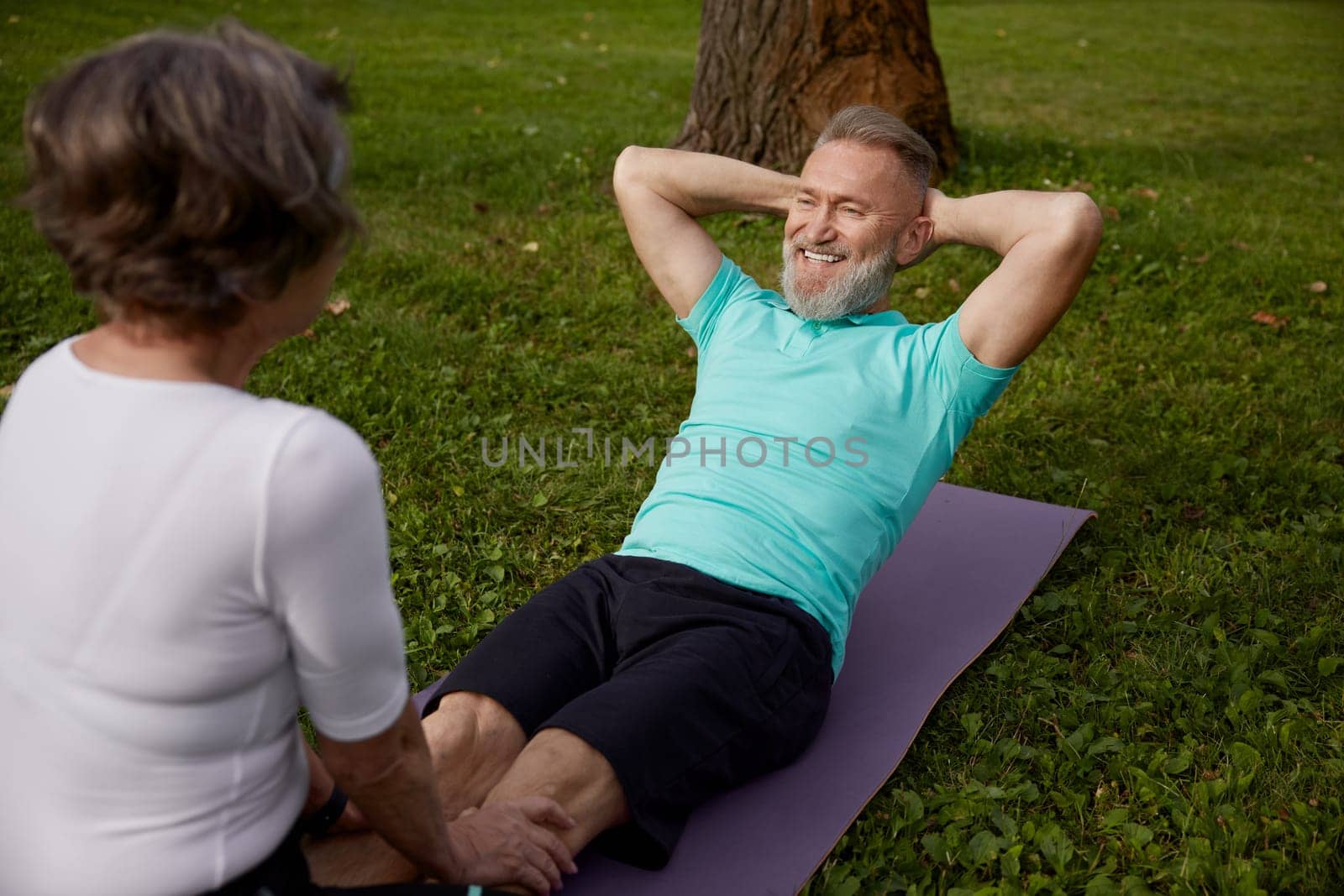 Happy married couple of retirees doing training workout in park. Elderly wife helping senior husband doing abdominal crunch. Sports leisure activity for pensioner