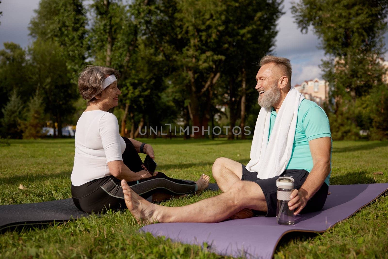 Senior couple sitting on fitness mats rest after yoga training outdoors in city park. Elderly woman using smartphone, mature man drinking water. Sports weekend and free time on retirement