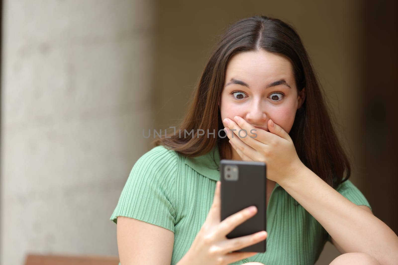 Front view portrait of a surprised woman checking cell phone news in the street