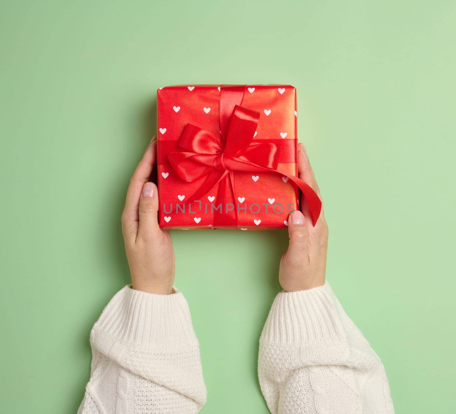 Female hands holding a gift box with a red ribbon on a green background. View from above