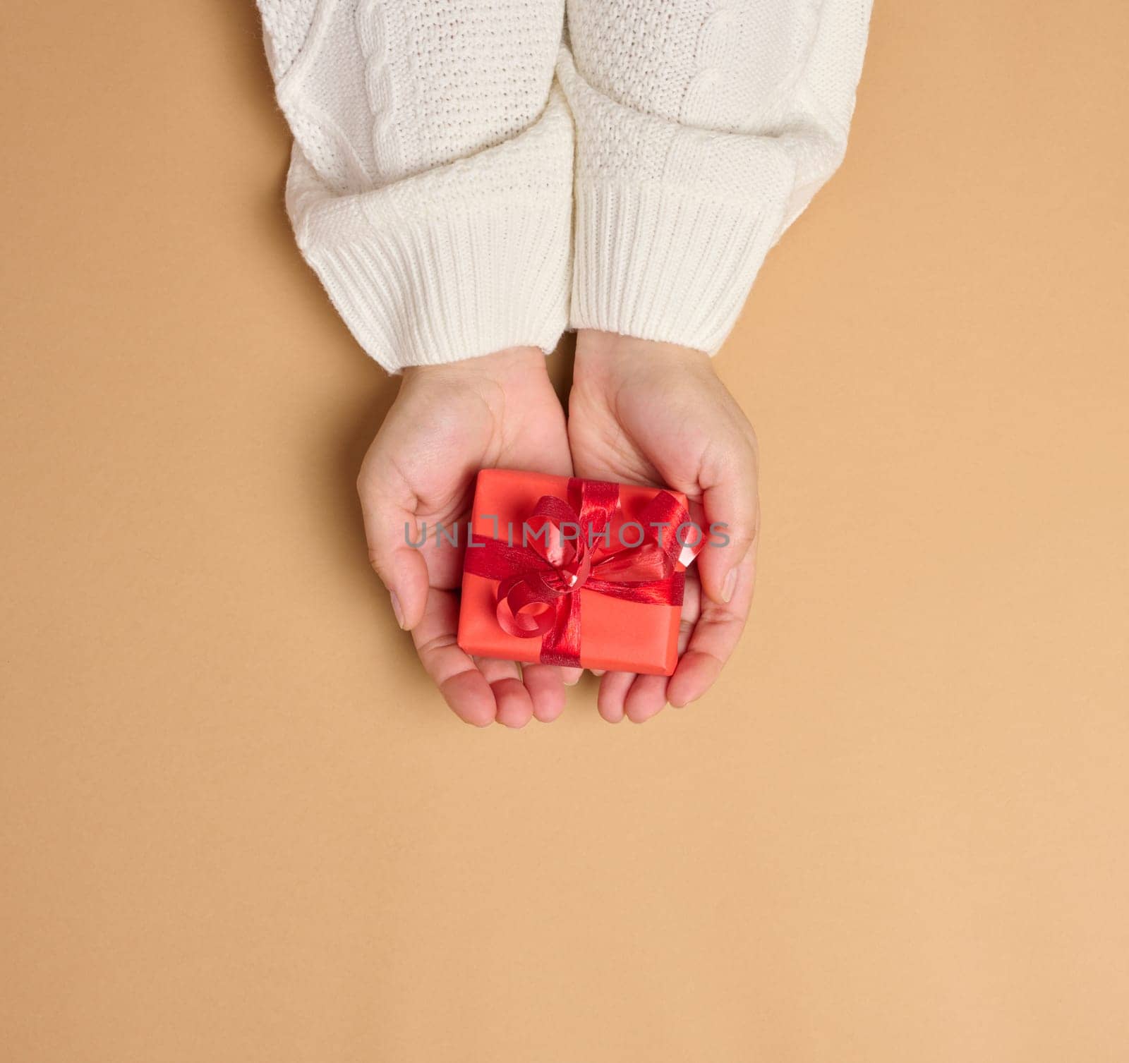 Two female hands holding a red box tied with a ribbon on a brown background, top view