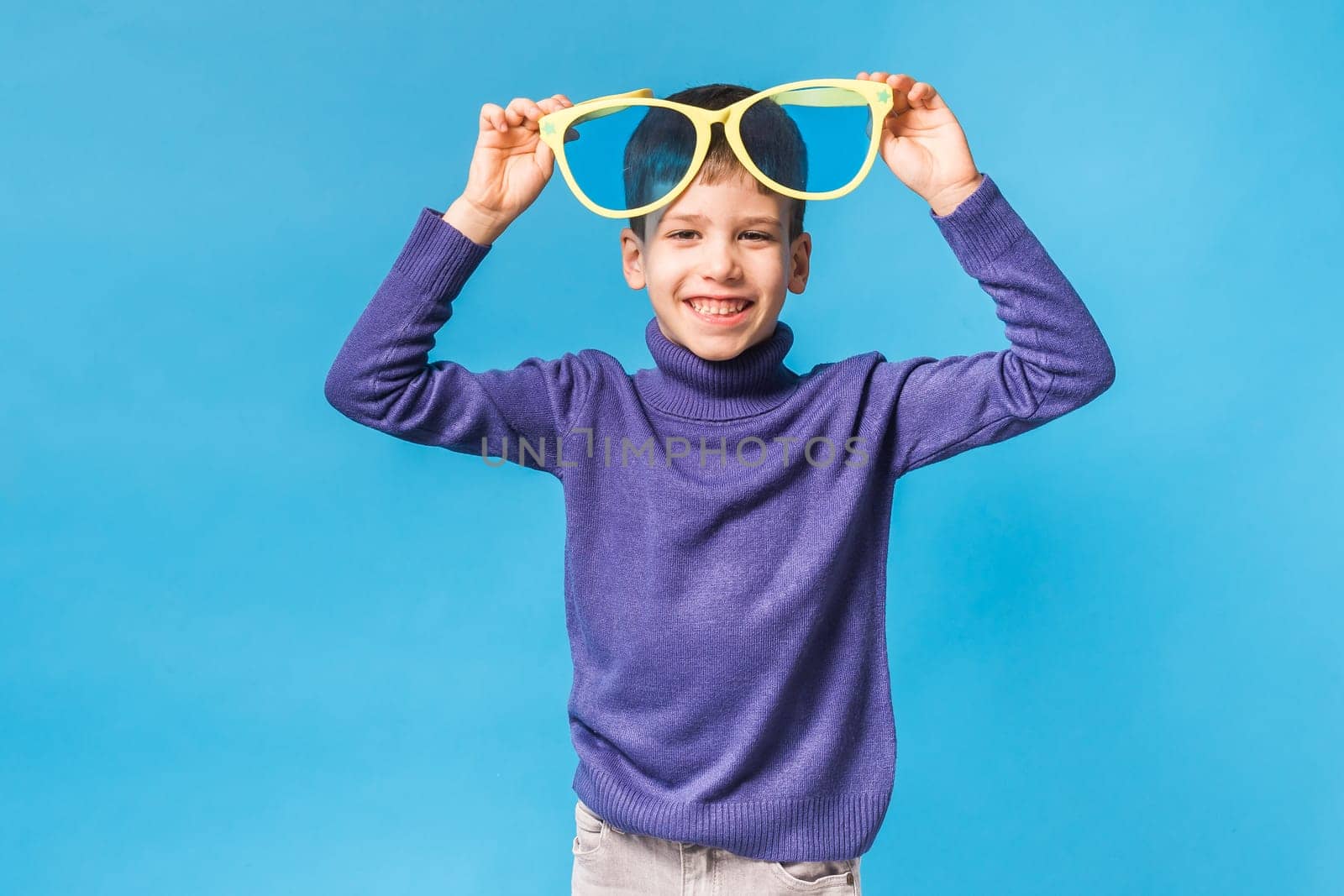 Cheerful little boy generation alpha in big glasses express a surprised face holds mockup board isolated on blue background with copy space.