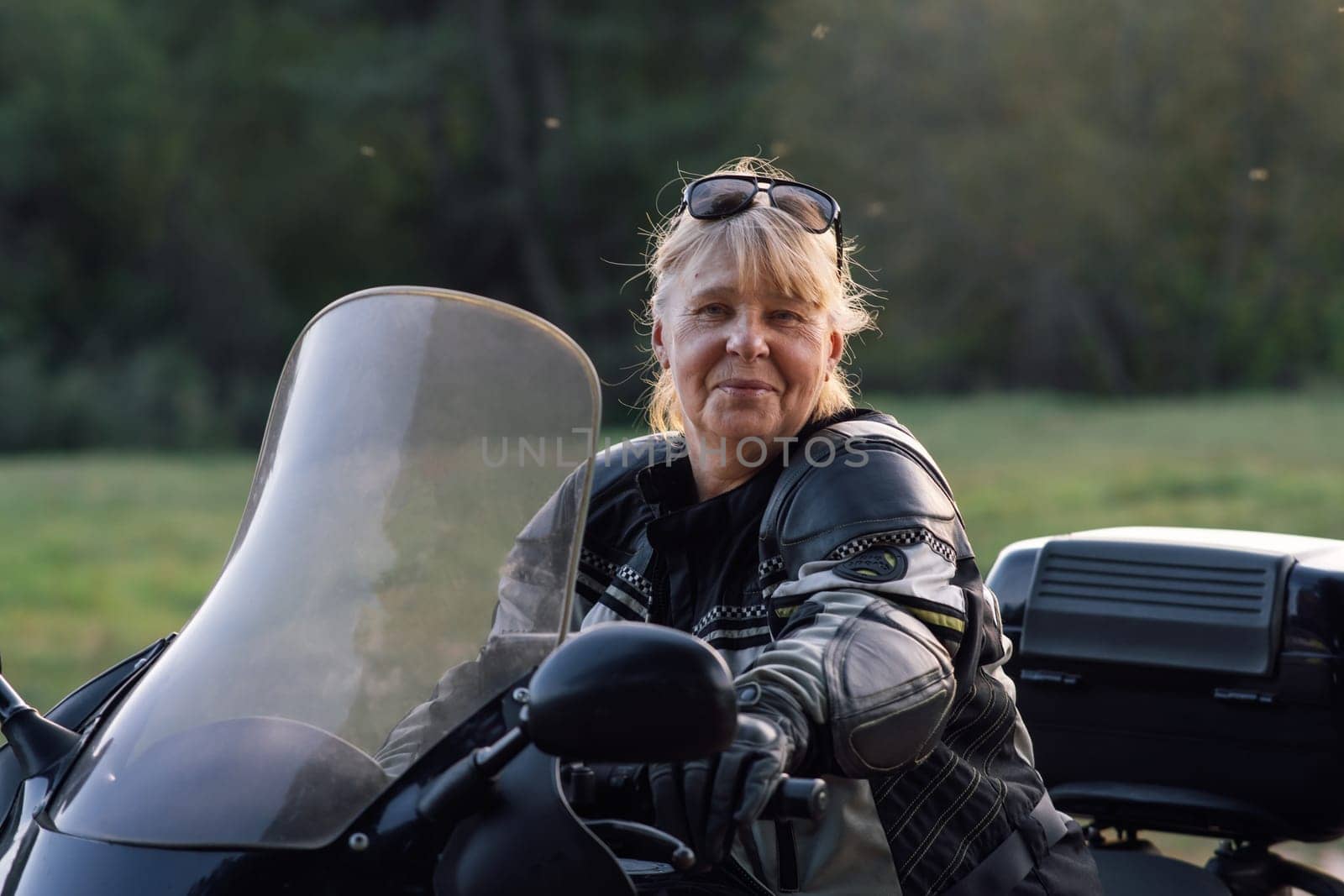 Happy smiling blonde middle-aged motorcyclist in leather jacket, black gloves is sitting on her bike on autumn day. Hobby . Healthy . Emotional relief. Excellent physical shape.Tourism and travel.