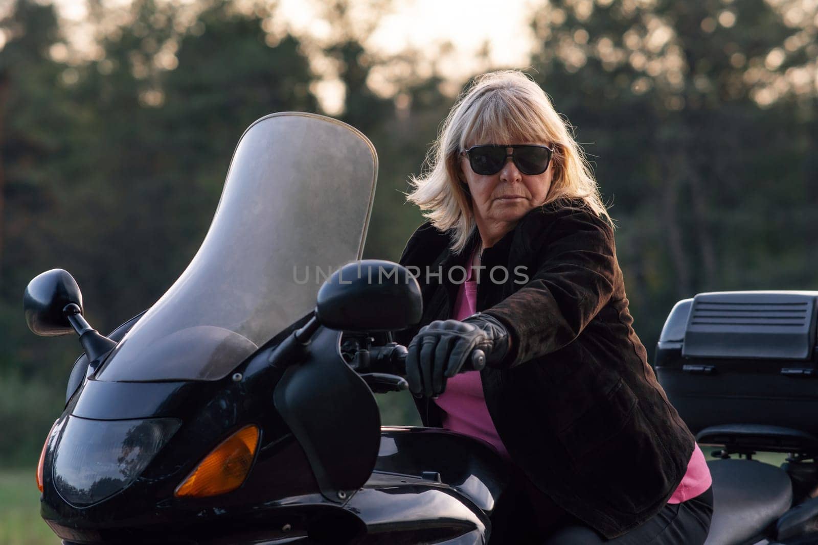 Stylish blonde motorcycle tourist, super mom, grandmother in black gloves and black glasses, sitting on her bike. Hobby motorcycle tourism. Healthy lifestyle. Excellent physical shape.Tourism travel.