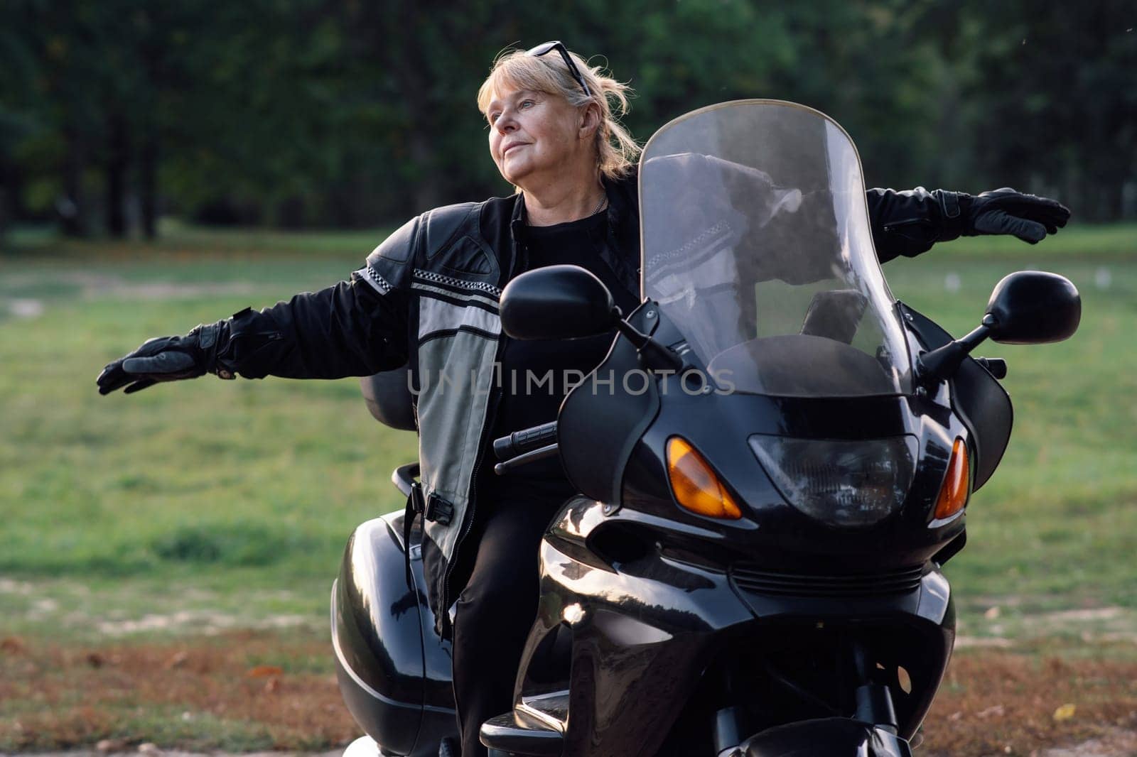 Towards adventures. Happy mature blonde motorcyclist has her arms outstretched, sitting on her black bike. Hobby motorcycle tourism. Emotional relief. Excellent physical shape. Tourism and travel.