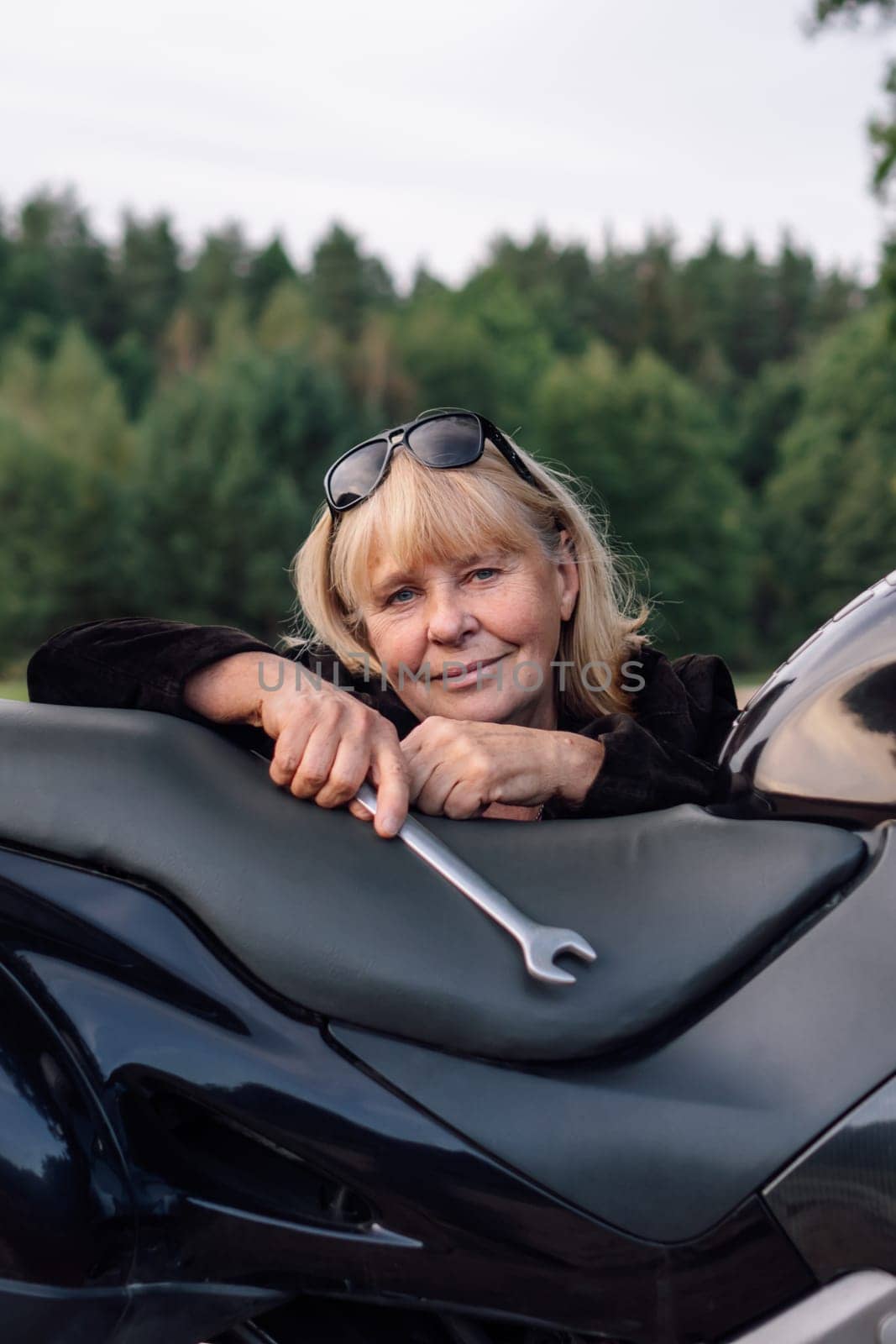 Vertical portrait of mature blonde woman with wrench on her black motorcycle in nature on autumn day. Hobby motorcycle tourism.Maintenance of motorcycles . Female mechanic. Tourism and travel.