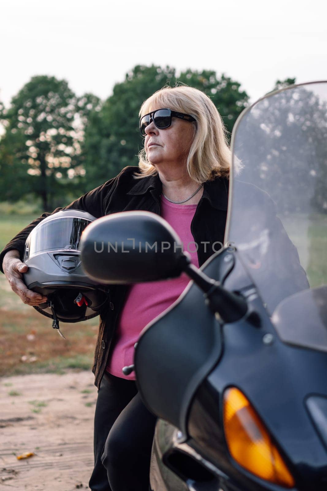 Vertical portrait of mature blonde in jacket with helmet in her hands standing near motorcycle in autumn in nature. Motorcycle tourism. Healthy lifestyle. Sport and strength of character.
