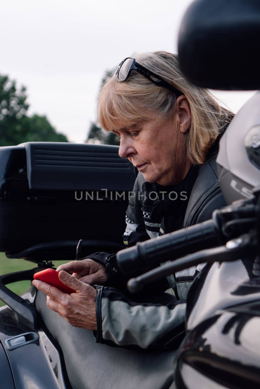 Communication in travel. Mature blonde on black motorcycle, holding mobile phone in nature during stop. Hobby - motorcycle tourism. Female biker. Tourism and travel. Strong independent women.
