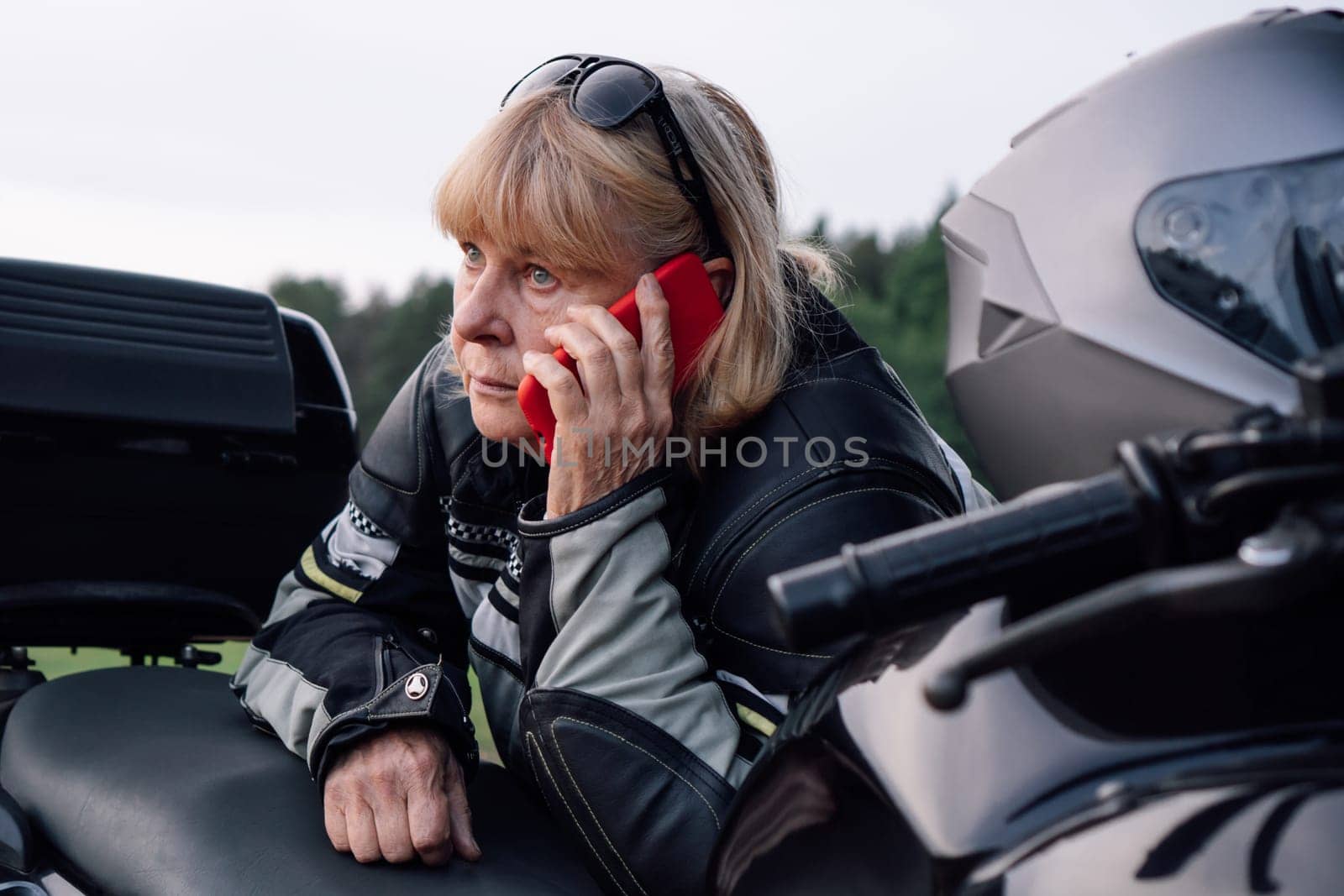 Tired middle-aged biker woman is talking on red phone about her black motorcycle on autumn day on road. Hobby - motorcycle tourism. Female biker. Communication in travel. Mobile communications.