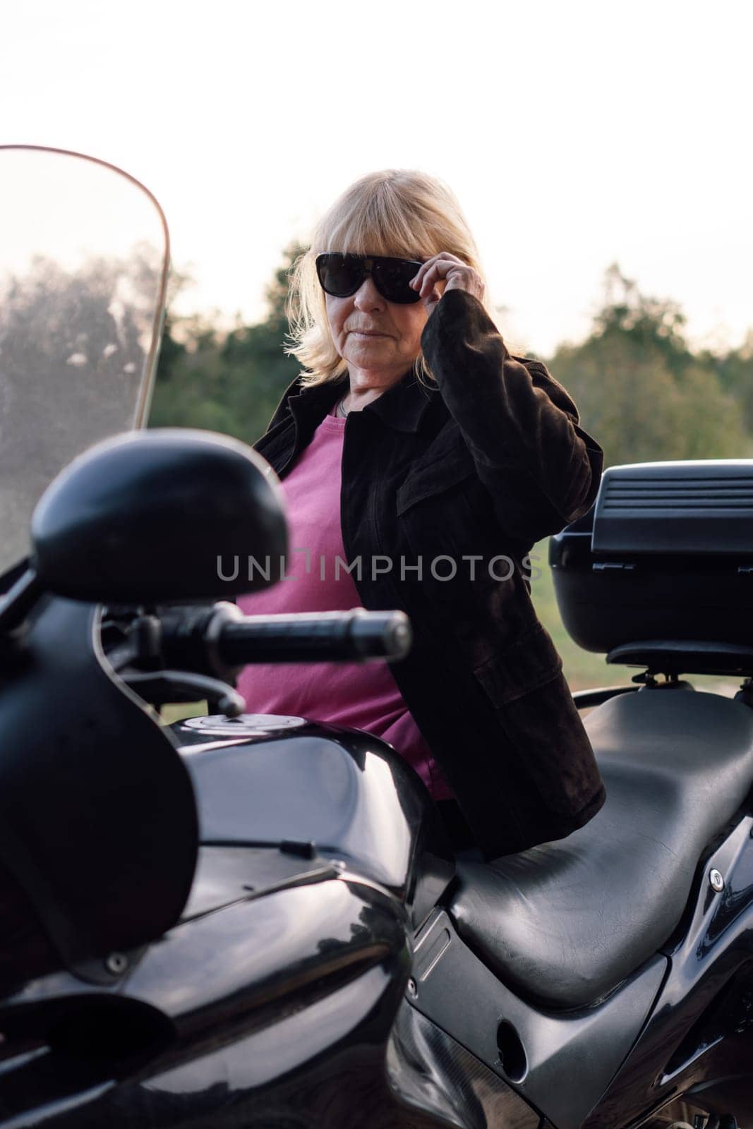 Portrait of mature blonde motorcyclist in black glasses in leather jacket, sitting on her bike on autumn day. Hobby . Healthy lifestyle. Emotional relief. Excellent physical shape. Tourism travel.