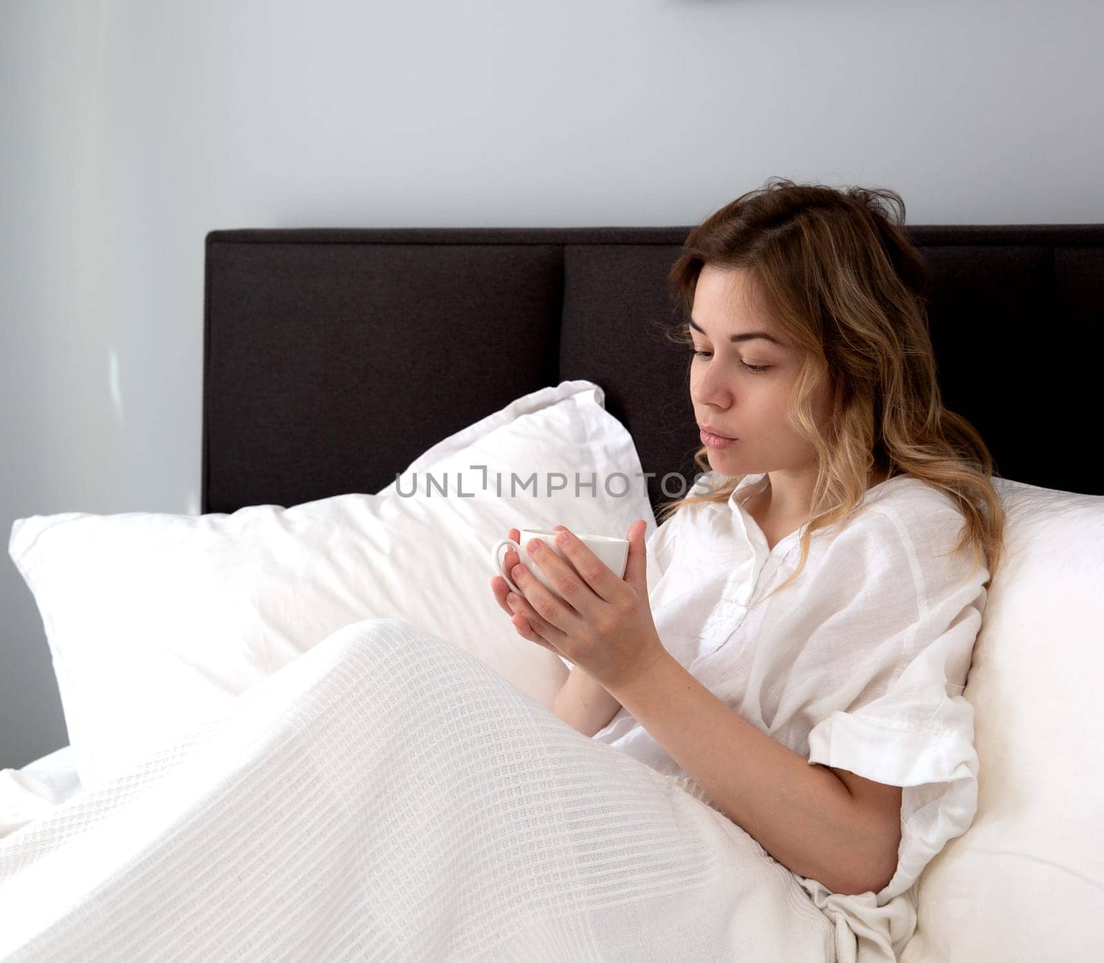 Beautiful blonde girl with a white cup sitting in a white bed in the morning light. High quality photo