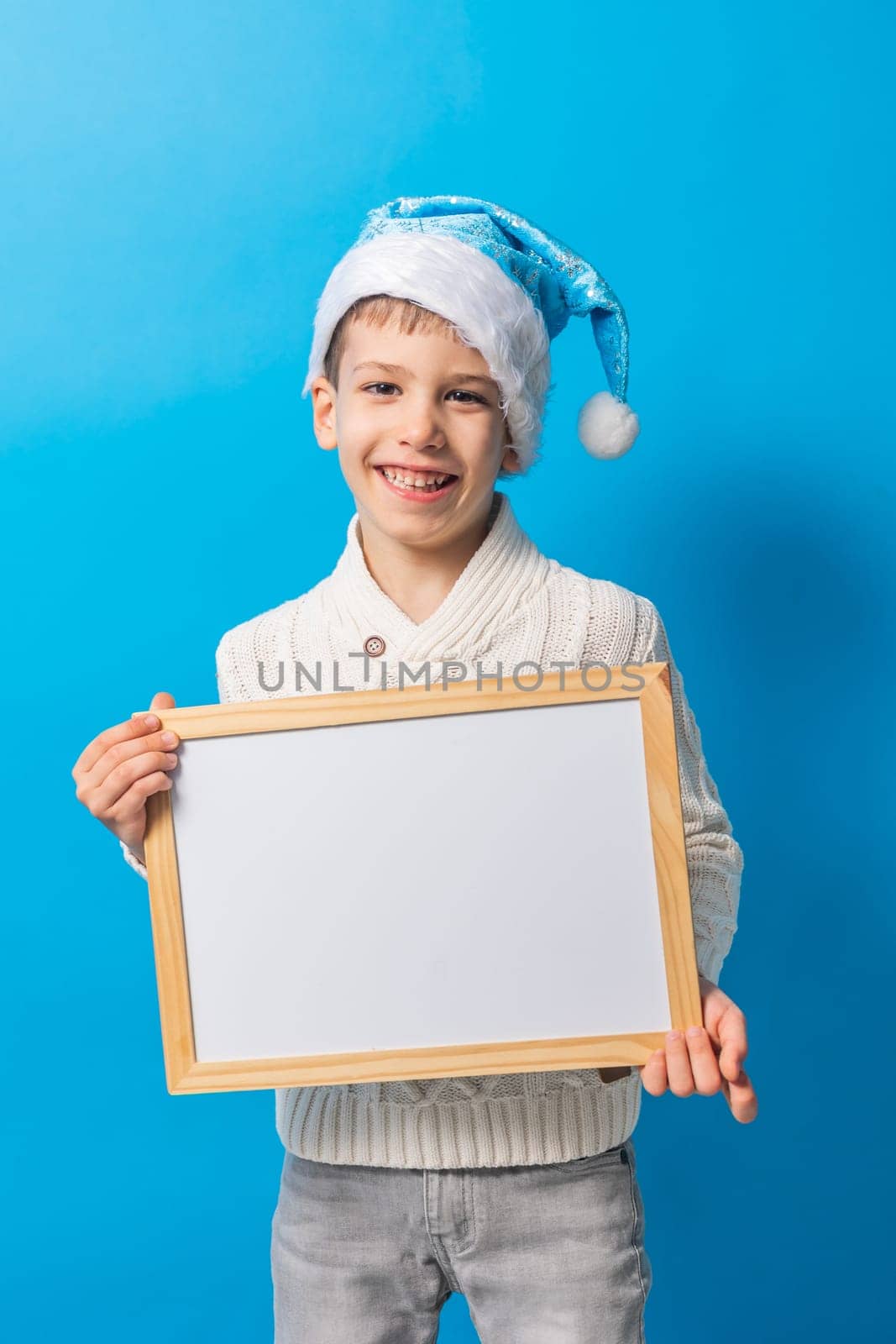 Child boy in blue santa hat and blank canvas. Copy space on canvas board for image or message. Kid in Christmas hat holds mockup poster and standing over blue background. Generation alpha and gen alpha.
