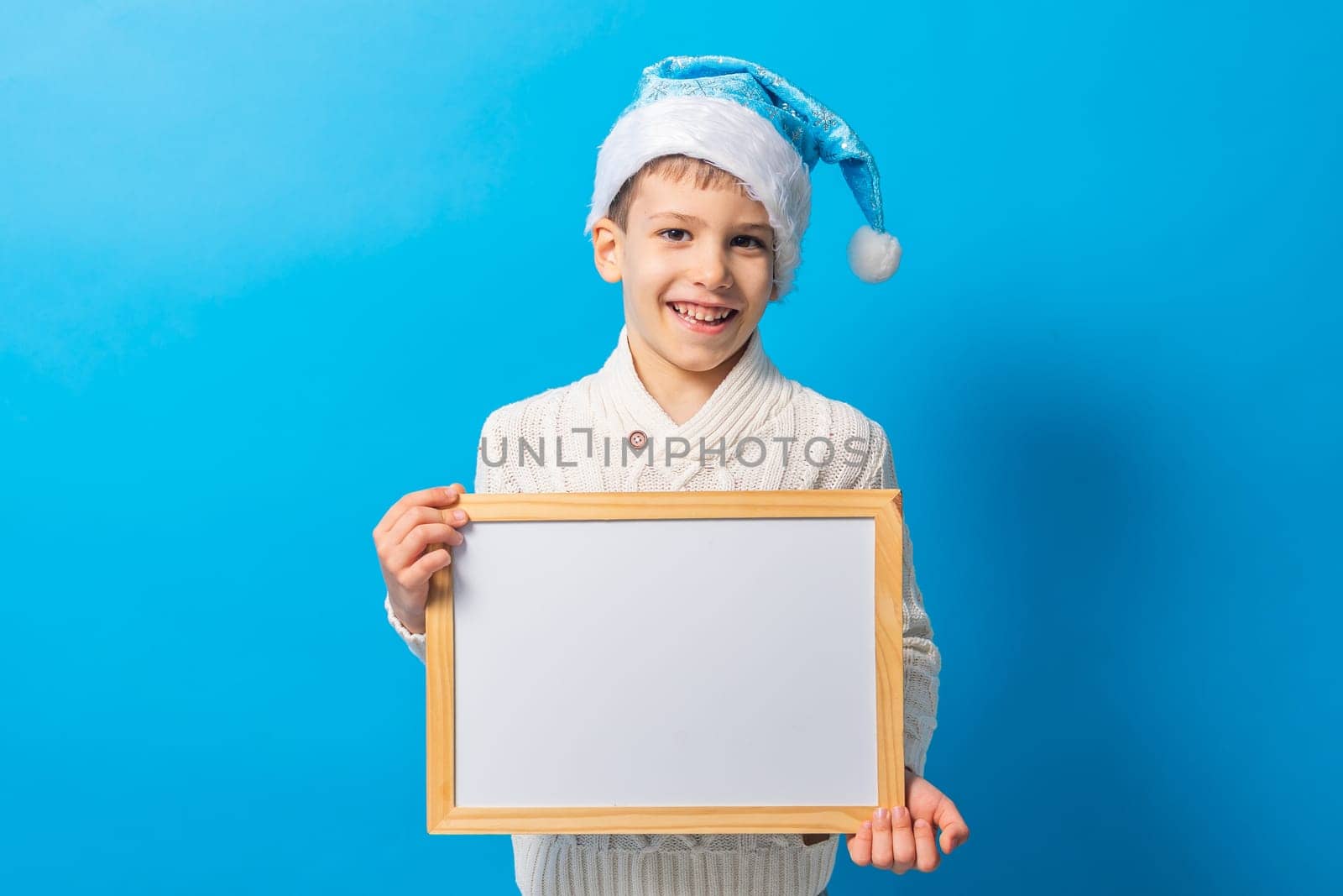 Child boy in blue santa hat and blank canvas. Copy space on canvas board for image or message. Kid in Christmas hat holds mockup poster and standing over blue background. Generation alpha and gen alpha.