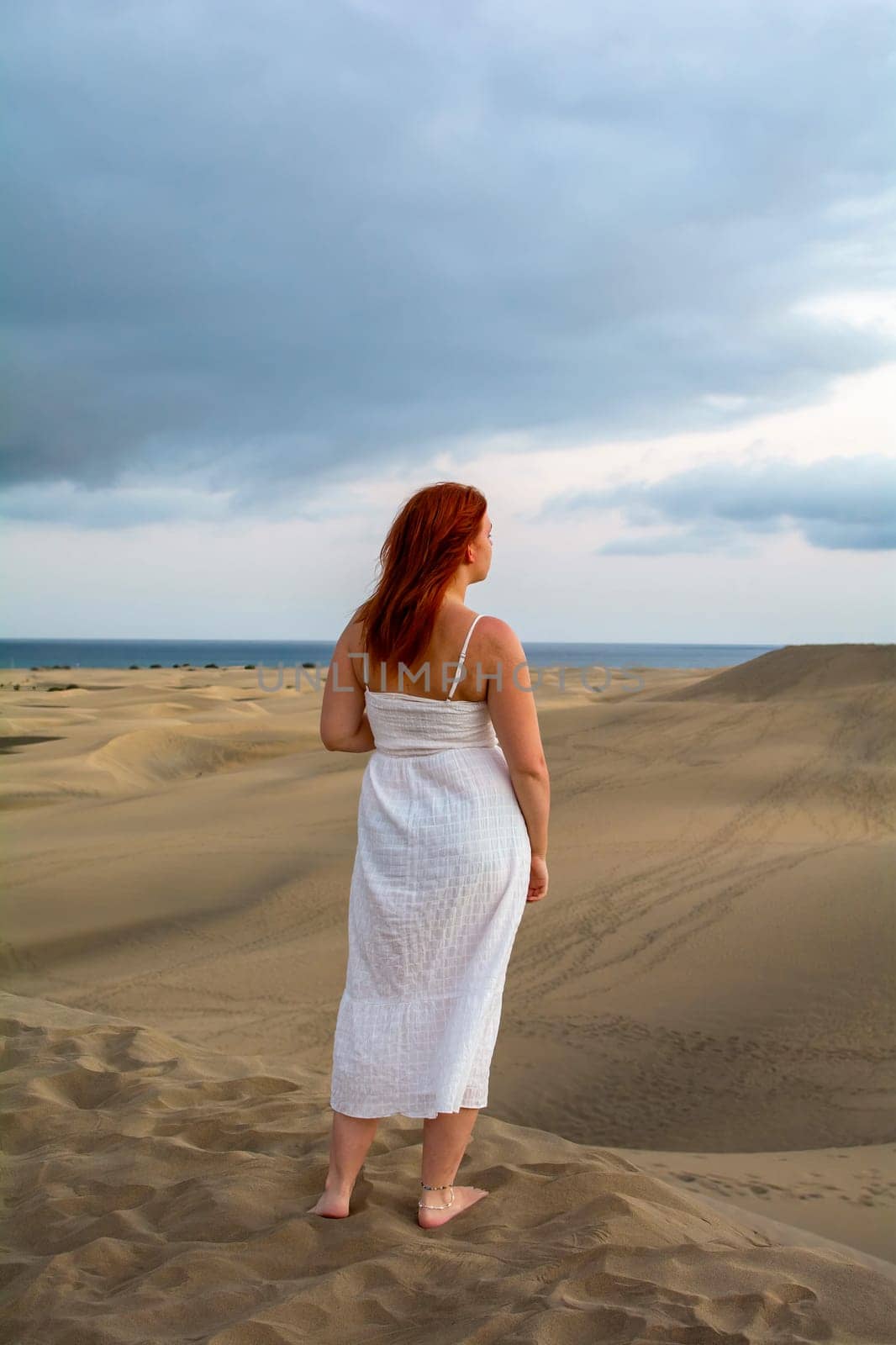 Young woman with red hair stands on a sand dune and looks at the sea