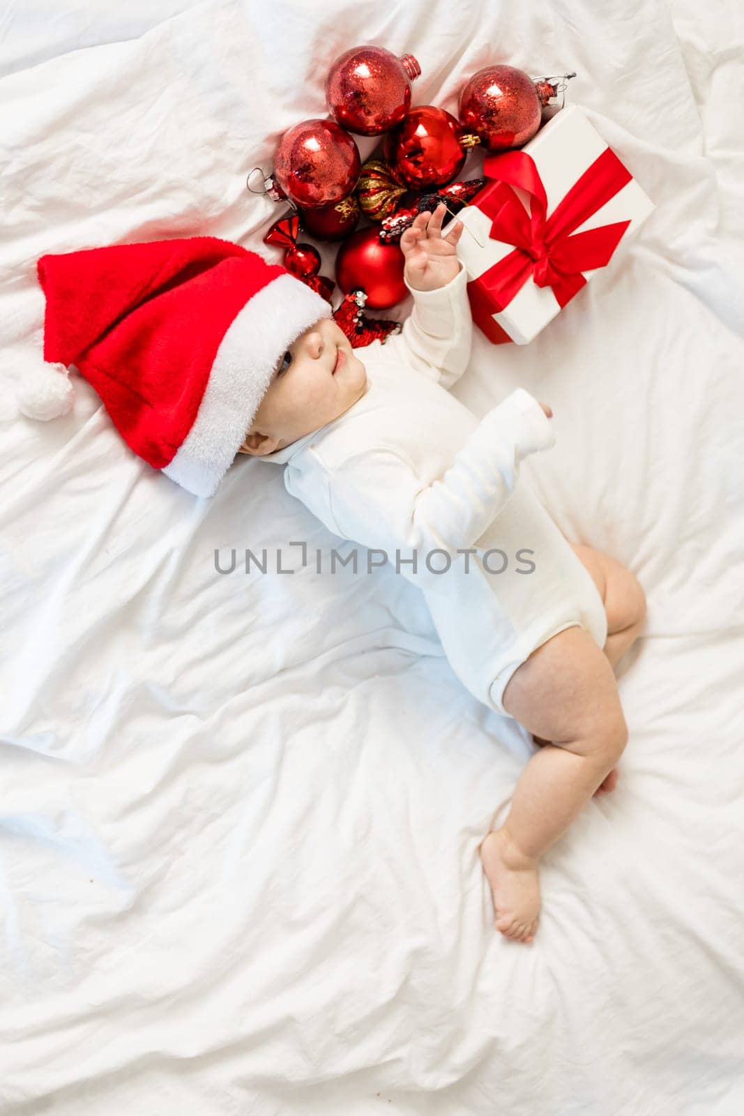 Christmas toddler in Santa hat lying on bed with gift box and festive baubles top view.