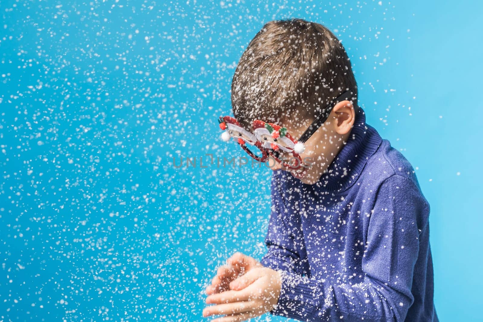 Child boy blowing snow out of his hands on blue background. Christmas and New Year holidays concept. Generation alpha and gen alpha children