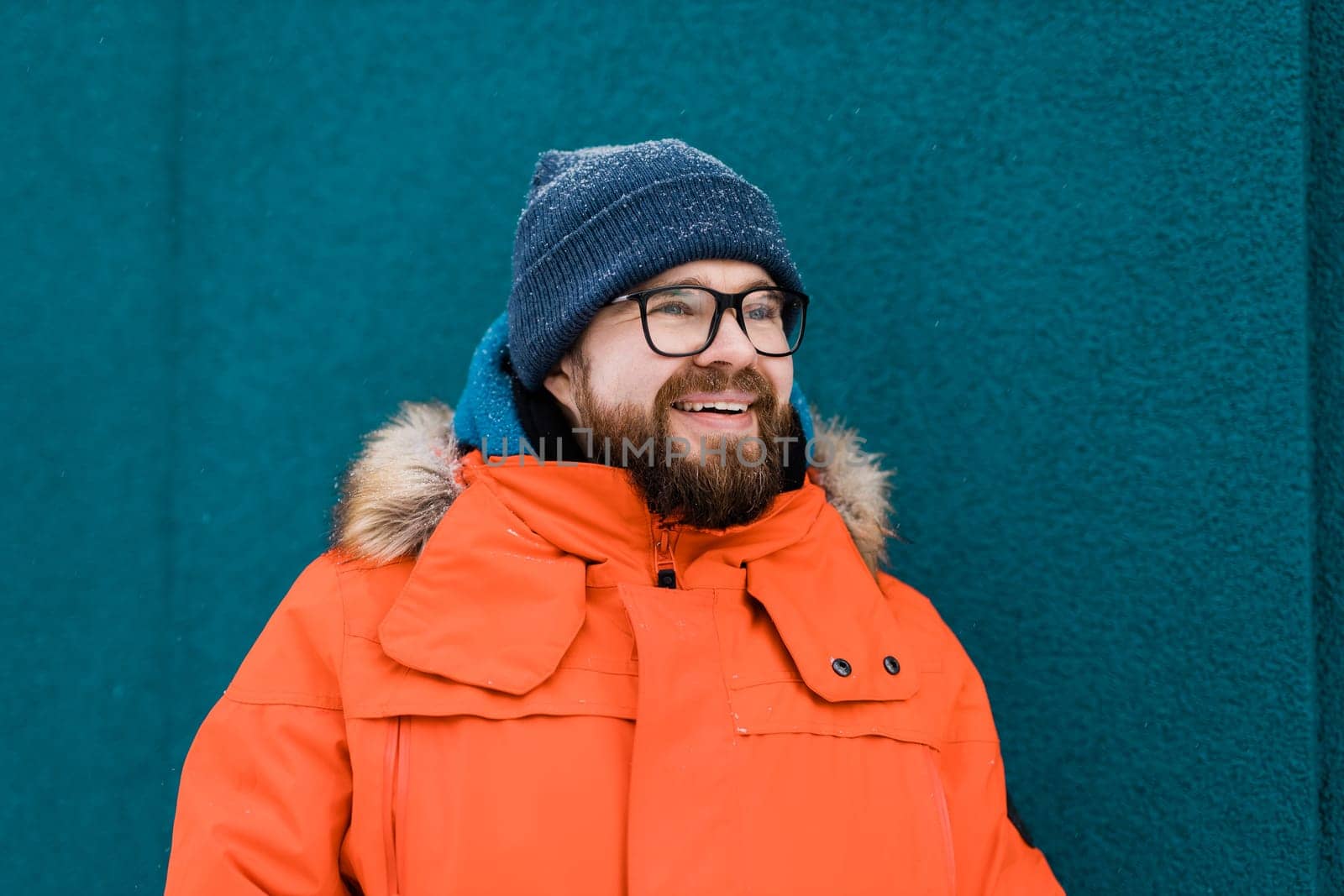 Portrait handsome eyeglasses young male smiling bearded stands on a blue wall background in a bright orange winter jacket with a hood with fur in winter. Millennial generation and Gen Y