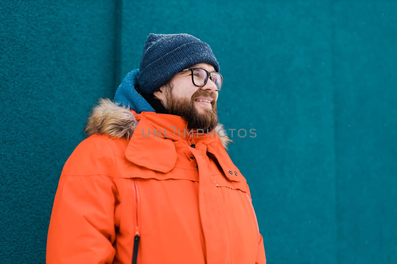 Portrait handsome eyeglasses young male smiling bearded stands on a blue wall background in a bright orange winter jacket with a hood with fur in winter. Millennial generation and Gen Y