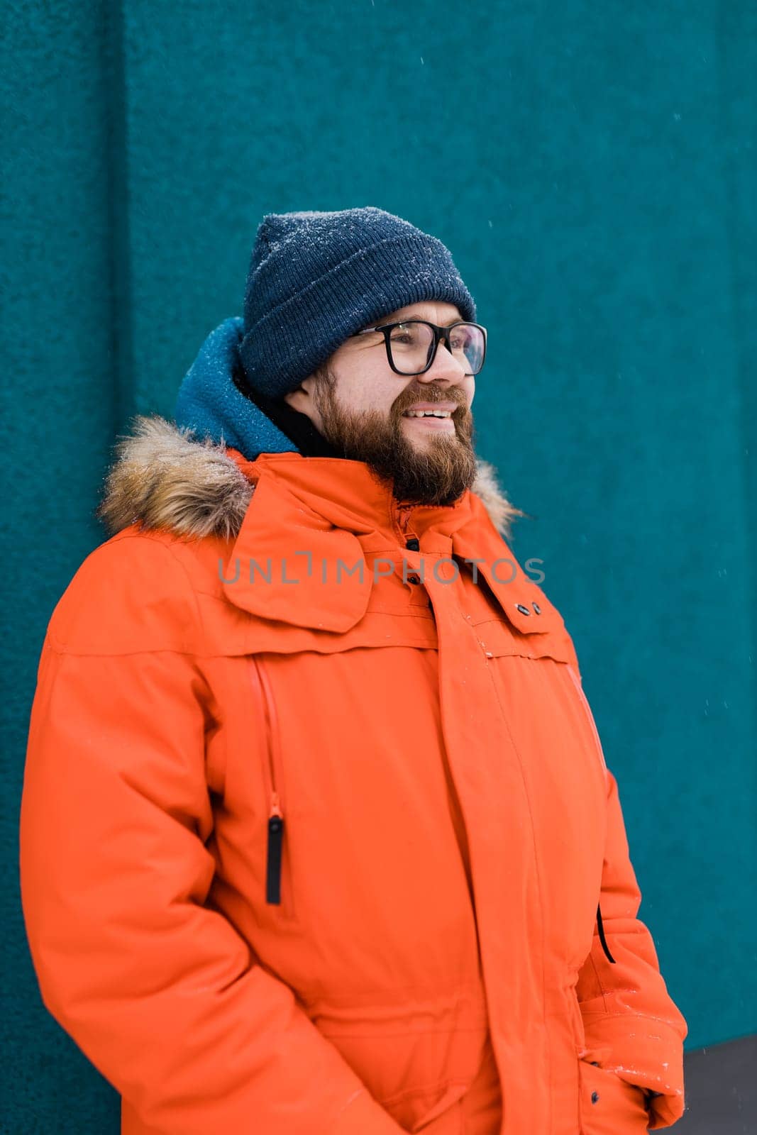 Portrait handsome eyeglasses young male smiling bearded stands on a blue wall background in a bright orange winter jacket with a hood with fur in winter. Millennial generation and Gen Y