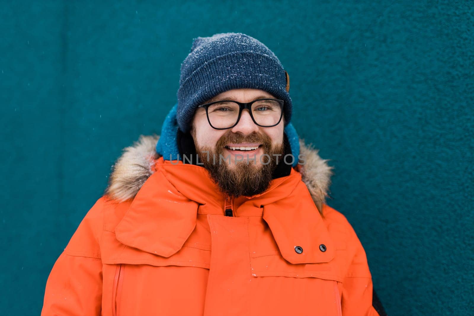 Portrait handsome eyeglasses young male smiling bearded stands on a blue wall background in a bright orange winter jacket with a hood with fur in winter. Millennial generation and Gen Y