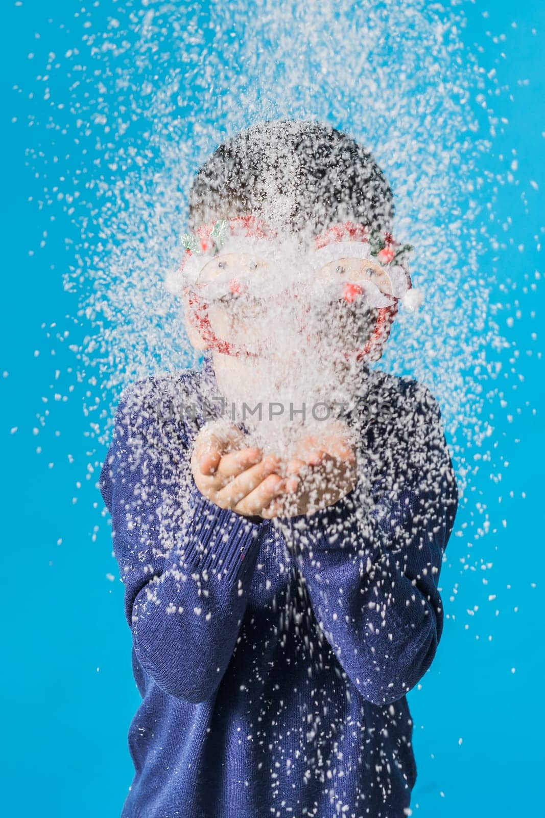 Child boy blowing snow out of his hands on blue background. Christmas and New Year holidays concept. Generation alpha and gen alpha children
