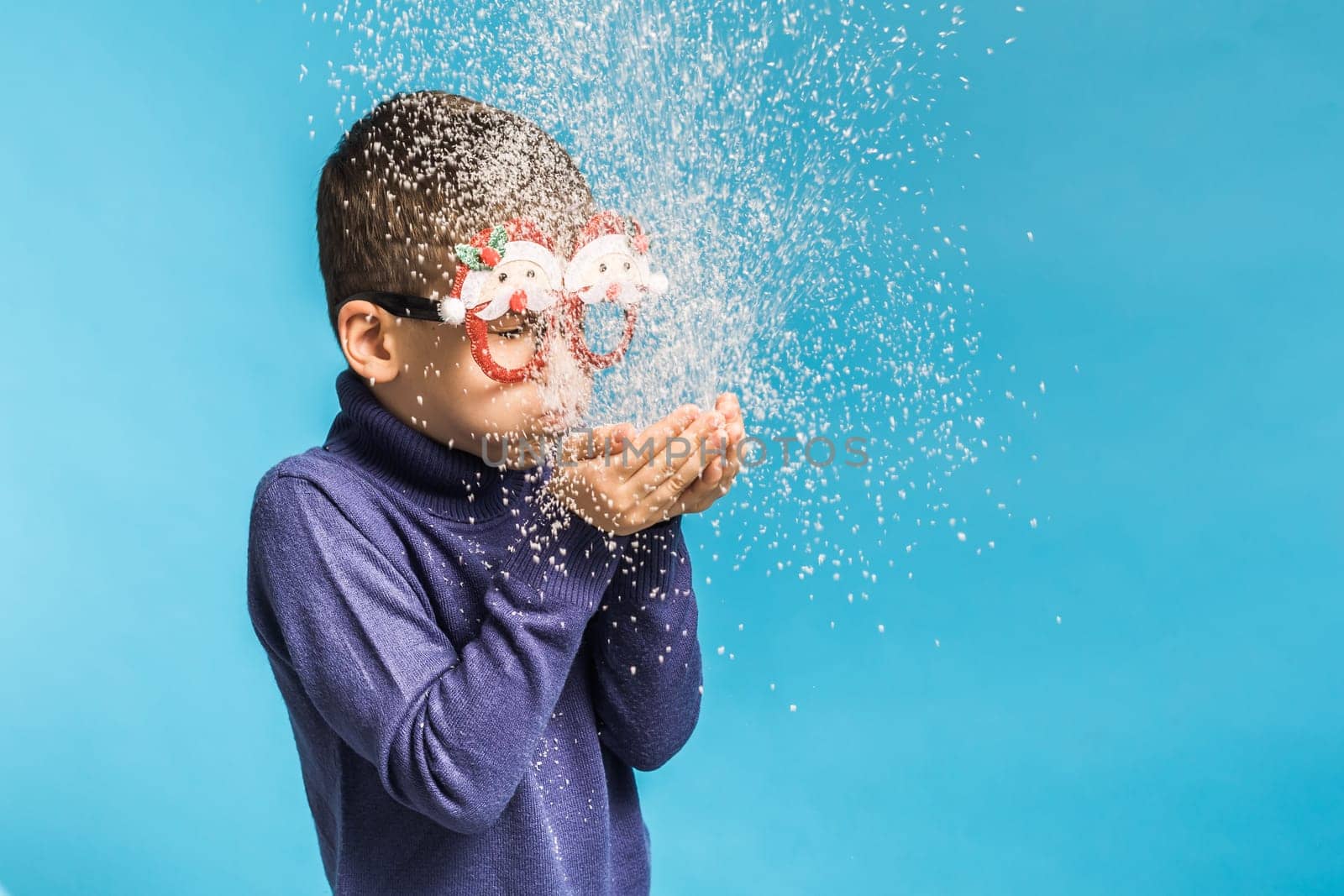 Child boy blowing snow out of his hands on blue background. Christmas and New Year holidays concept. Generation alpha and gen alpha children