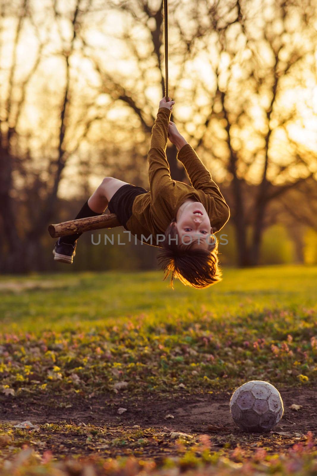 Active games outside in early autumn. Happy teenage boy is hanging upside down on homemade swing in nature, in park, in village in rays of sunset, football is lying nearby. Positive emotions.