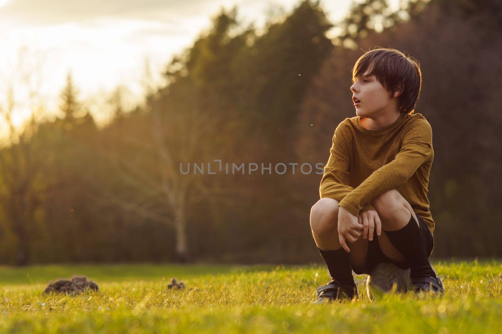 In early summer morning, teenager in long-sleeved T-shirt sits on soccer ball on field waiting for workout. Sports and health. Children's sports sections. Hobbies and hobbies. Summer sports camp.