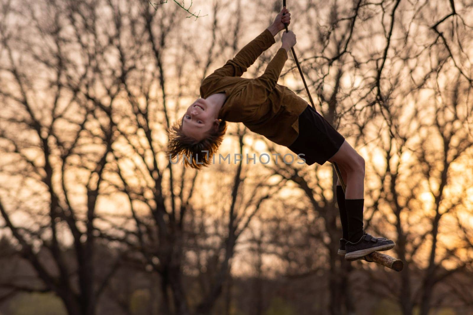 Happy teenage boy flies high up on homemade swing in nature, in park, in village against background of autumn trees in colors of sunset. Positive emotions. Child protection. Child safety. Recreation.