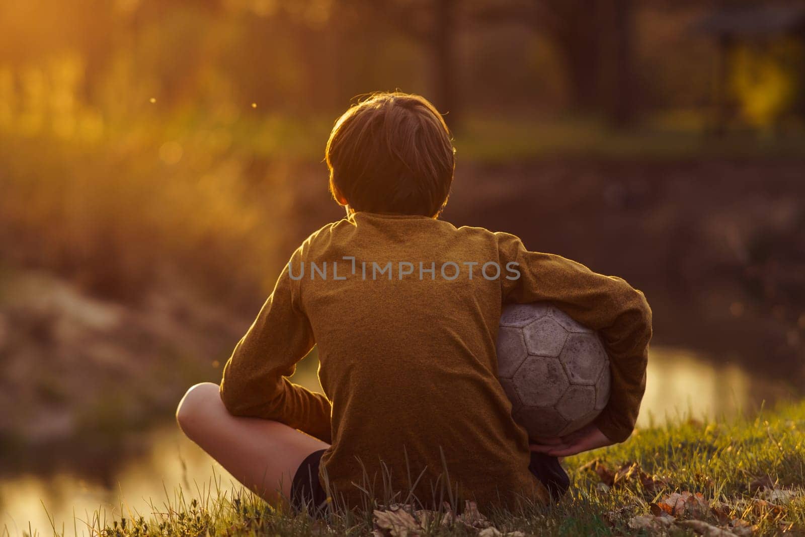 Teenager in long-sleeved T-shirt and ball under his arm sits with his back to camera after training on field and looks at stream. Children's sports sections. Football practice. Summer sports camp.