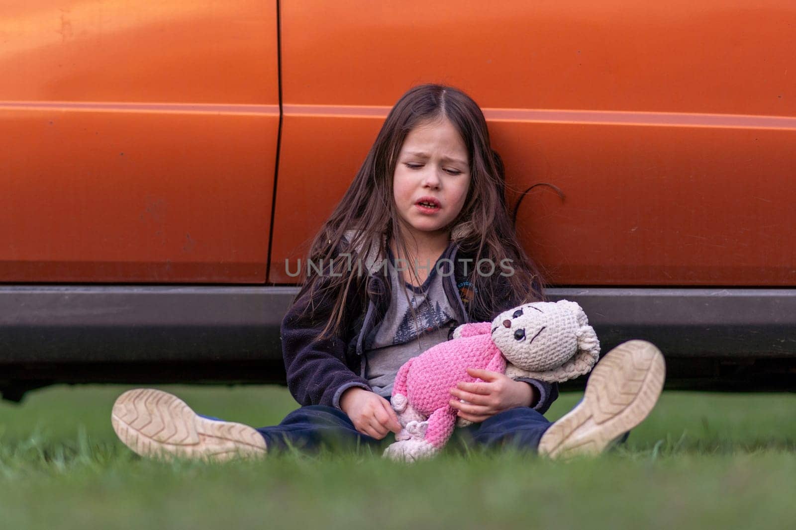 Children's problems. Little lonely girl is very upset, tired, holding toy in her hands, sitting on grass with her back pressed against car. Child is upset and offended. Inner world of child.
