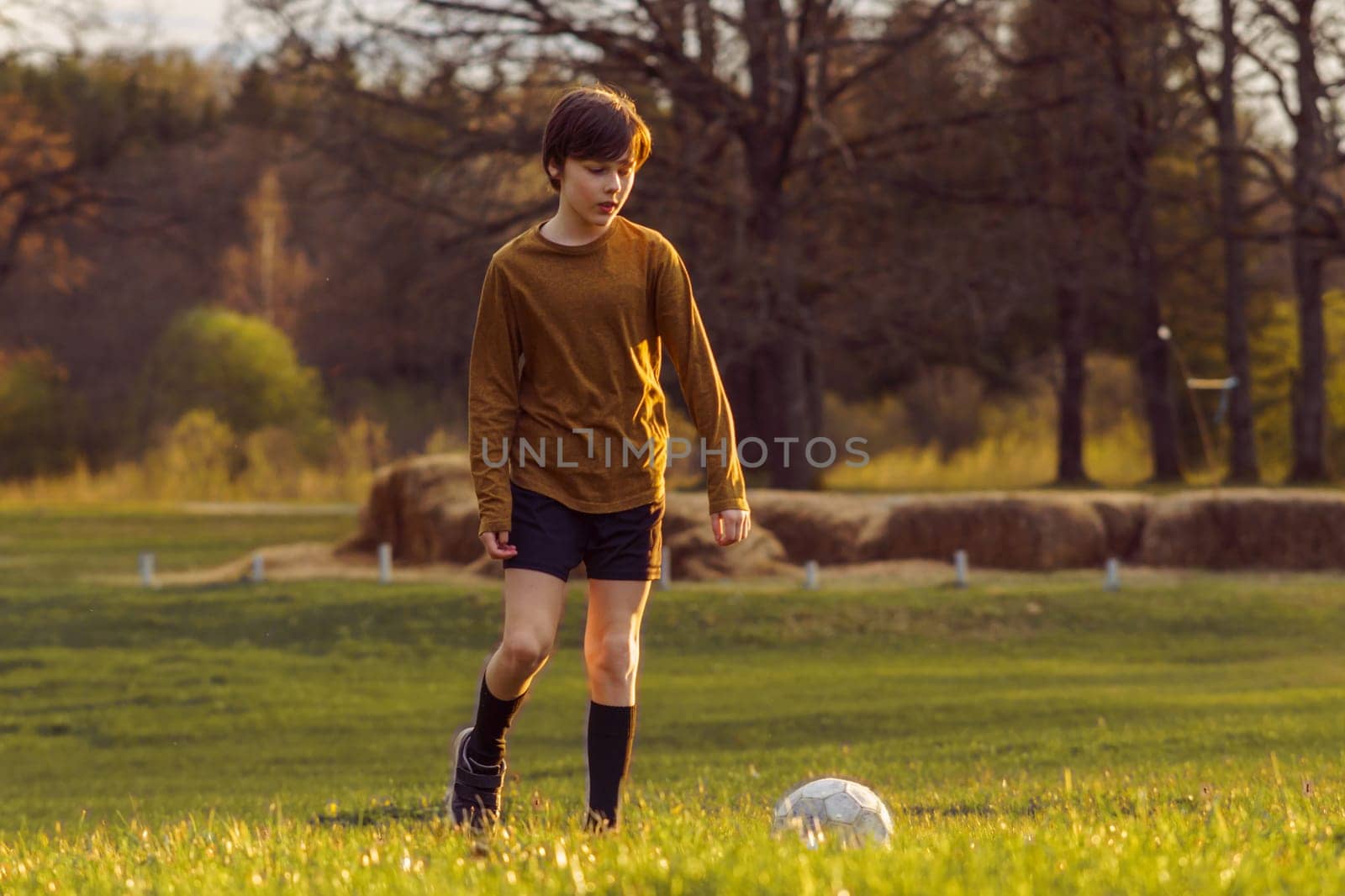 Teenage boy of 11-13 years old in sports uniform trains early in summer morning on field with soccer ball against background of stacks of mown grass. Sports and health. Sports activities .