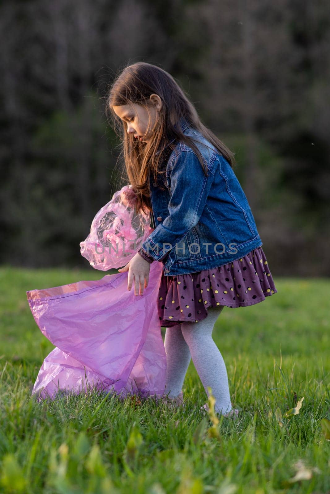 Vertical portrait of charming little girl with long hair in denim jacket collecting garbage in nature, in meadow on summer day. Environmental problems. We take care of nature. Saving planet.