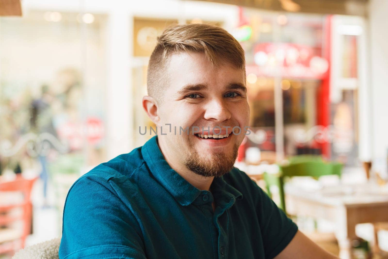 Close up of handsome Caucasian man with beard looking and smiling at camera with happy cheerful expression, sitting at restaurant, waiting for friends.