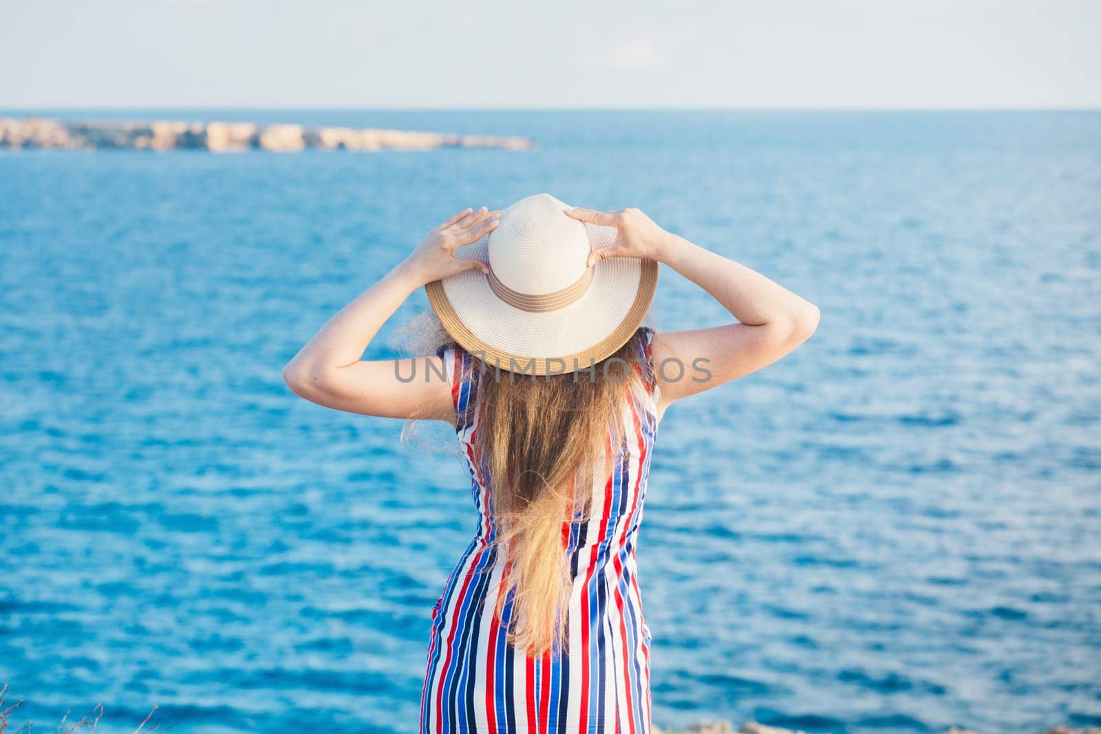 Closeup back view of woman sitting in white dress and hat looking out towards blue ocean and sky, isolated sea background