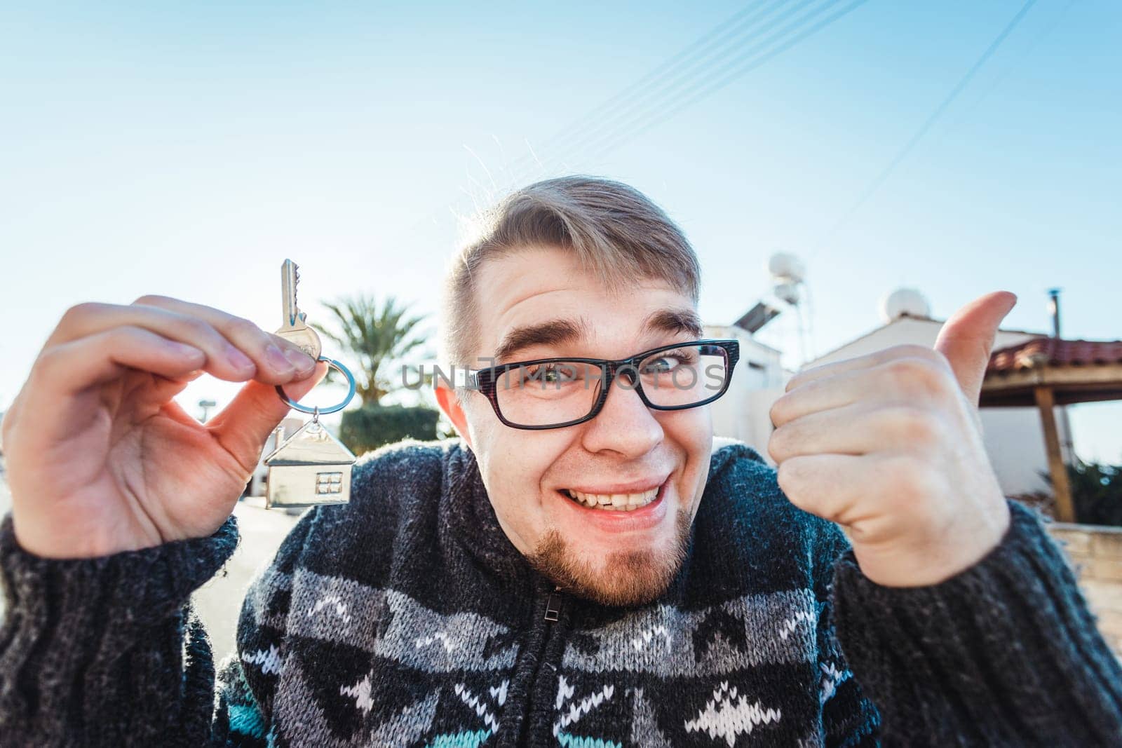Happy funny man holds house keys on house shaped keychain in front of a new home.