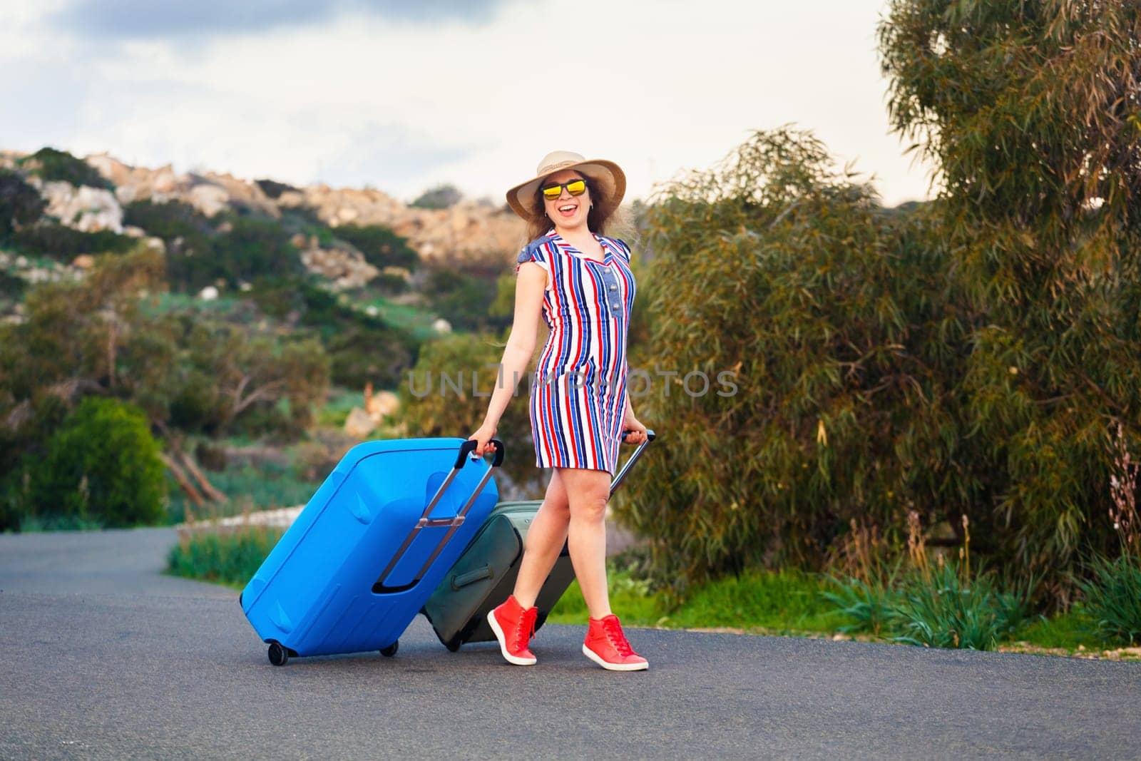 Traveler woman wearing a dress and hat sits on blue suitcase on the road and laughs .