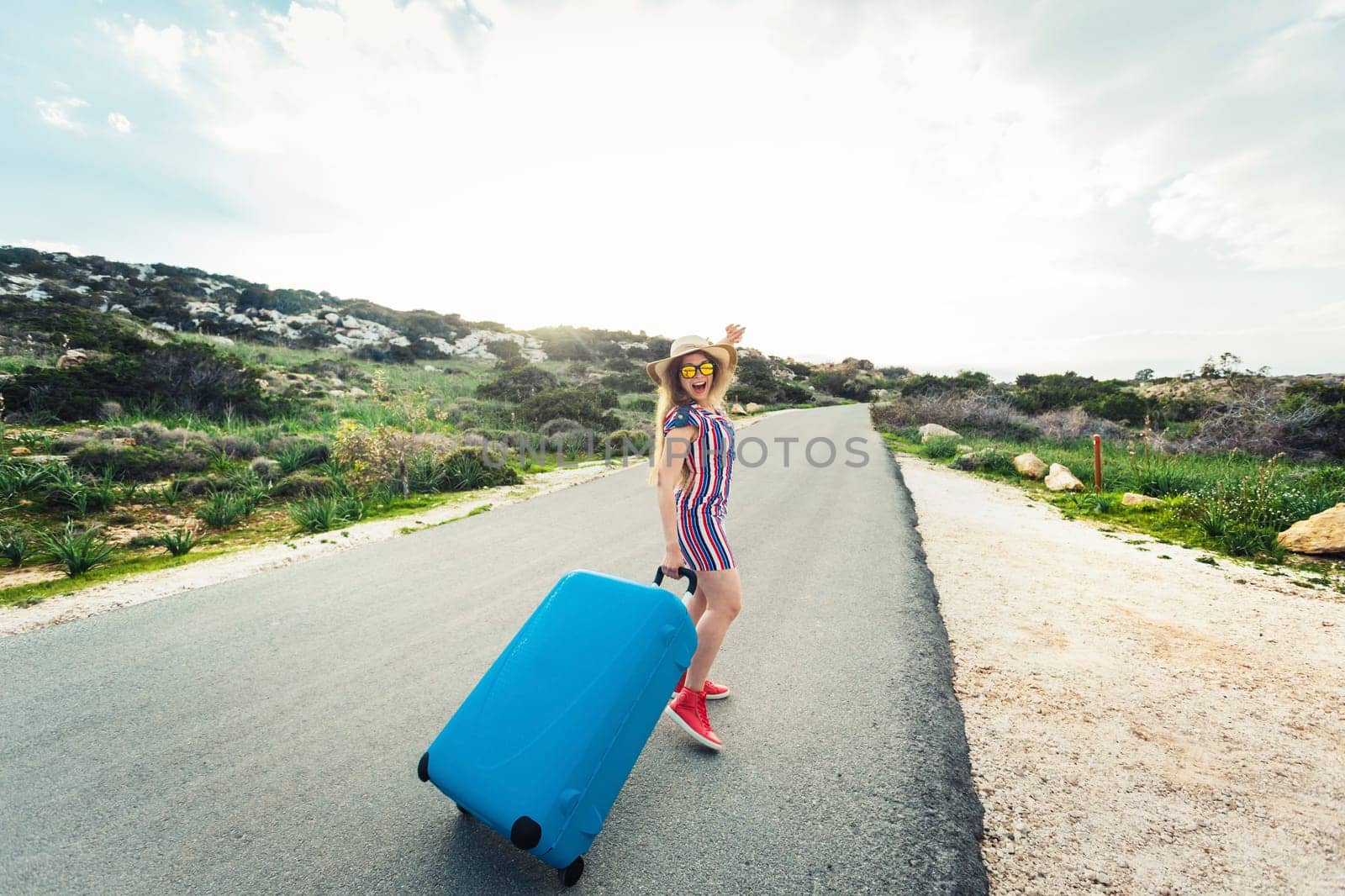 Traveler woman wearing a dress and hat sits on blue suitcase on the road and laughs .