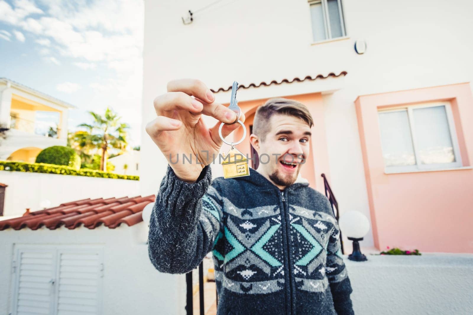 Happy funny man holds house keys on house shaped keychain in front of a new home.