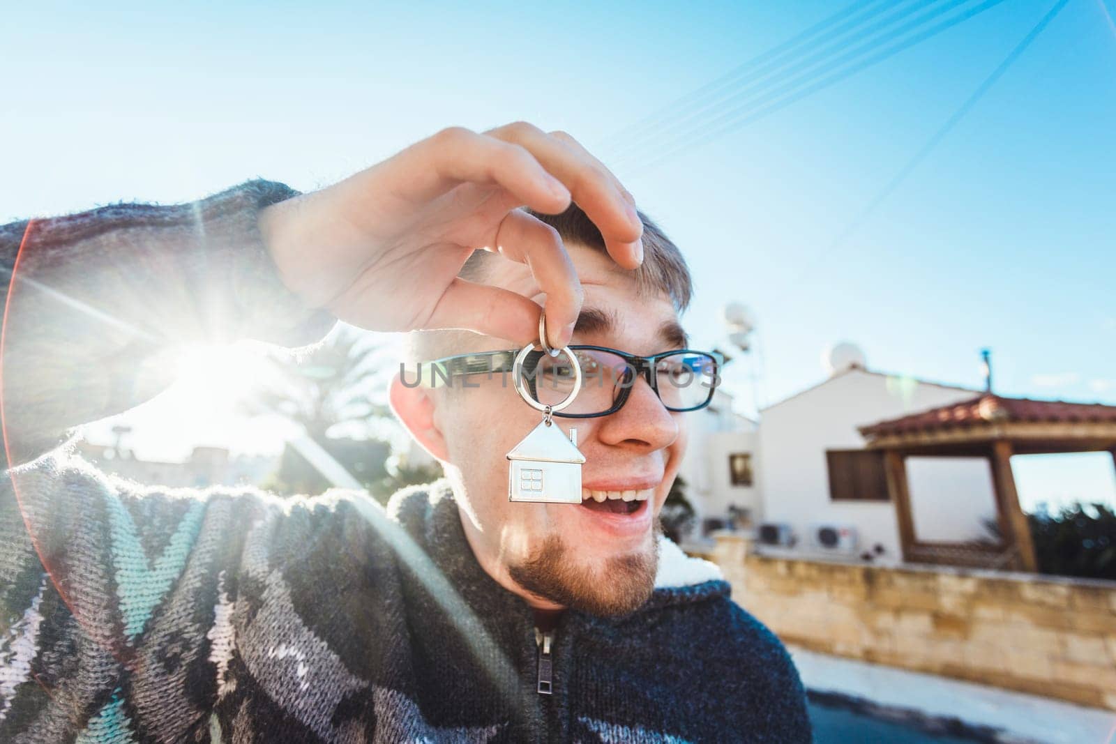 Happy funny man holds house keys on house shaped keychain in front of a new home.