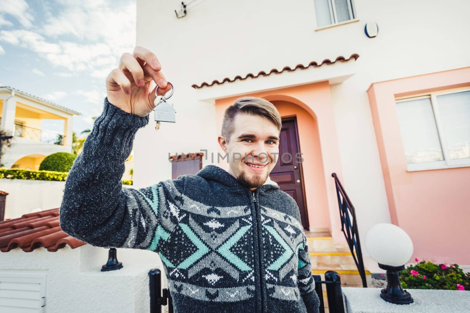 Happy funny man holds house keys on house shaped keychain in front of a new home.