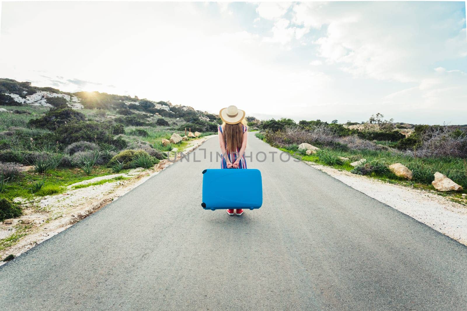 Back view of woman with blue suitcase on a road.