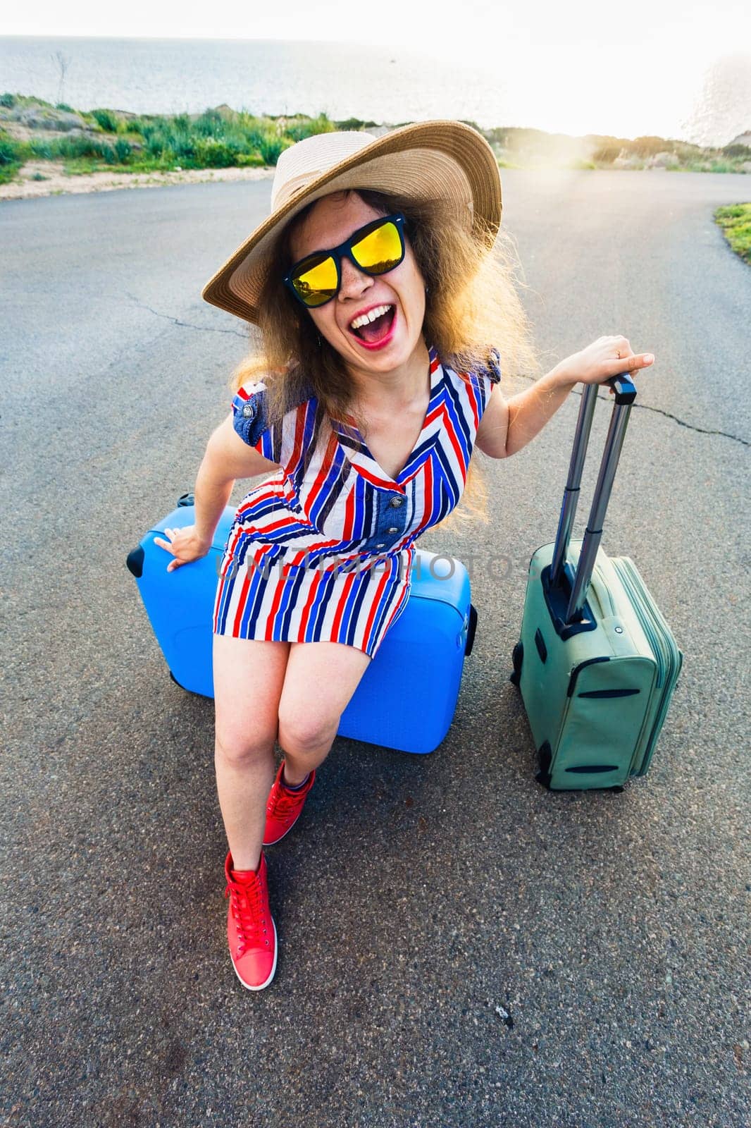 Happy traveler woman sitting on a suitcase on the road and smiling. Concept of travel, journey, trip.