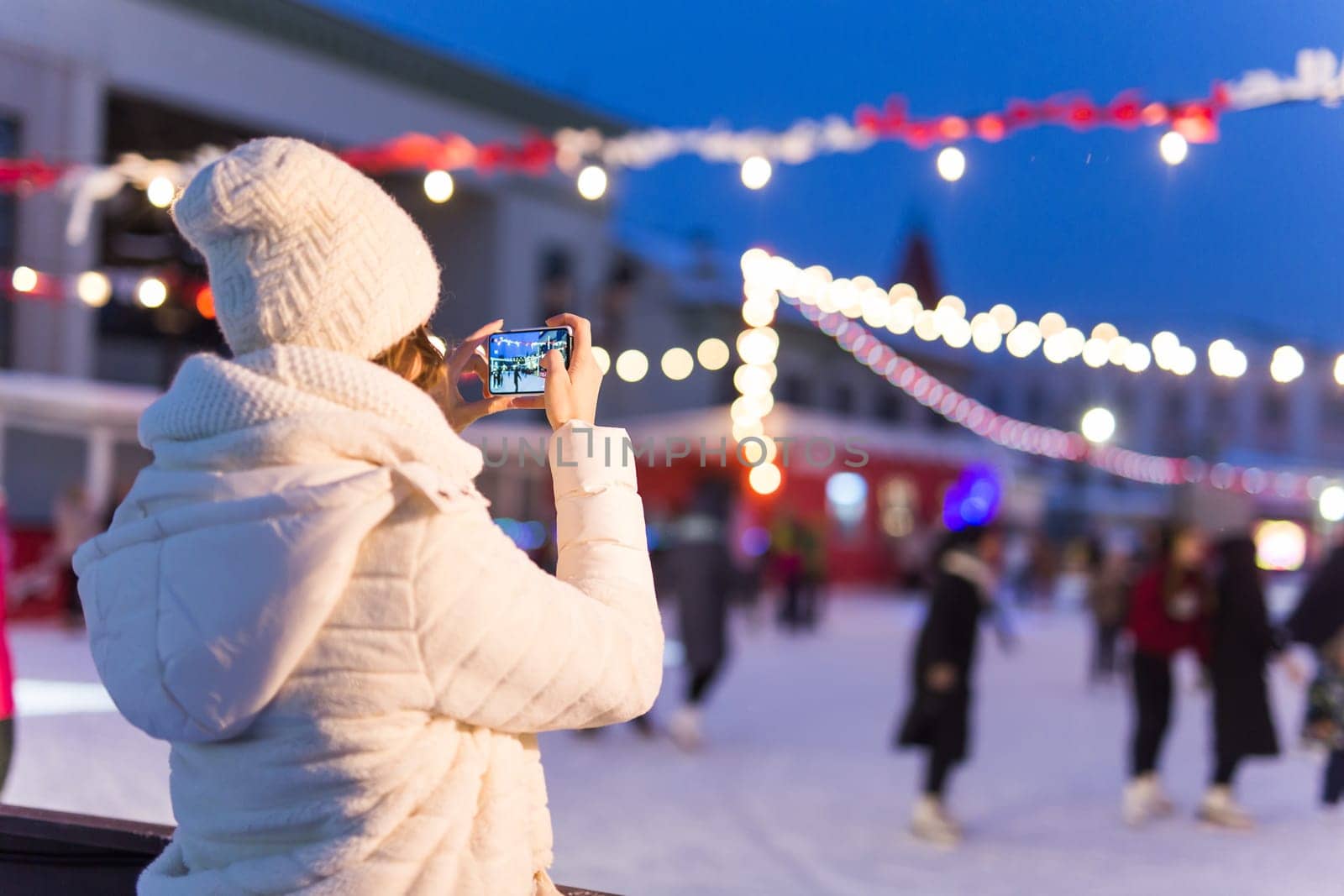 Christmas, winter, technology and leisure concept - happy young woman taking picture with smartphone on ice skating rink.