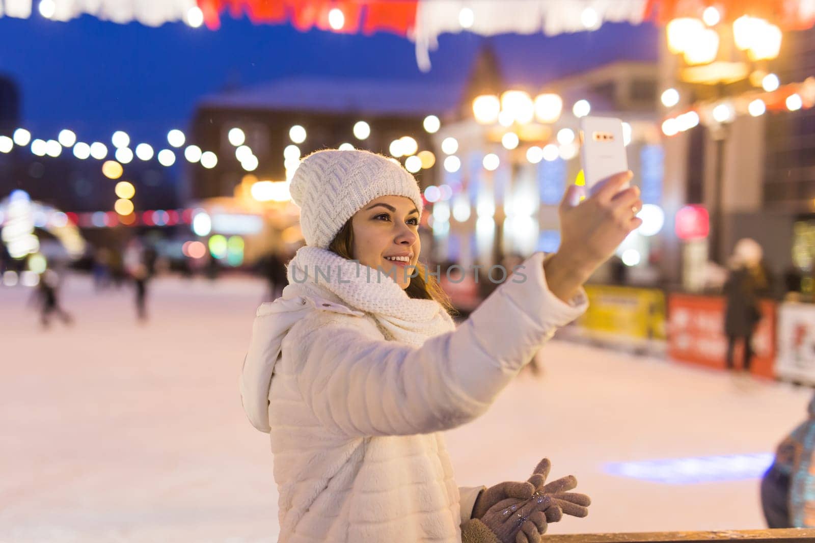 Christmas, winter, technology and leisure concept - happy young woman taking picture with smartphone on ice skating rink.