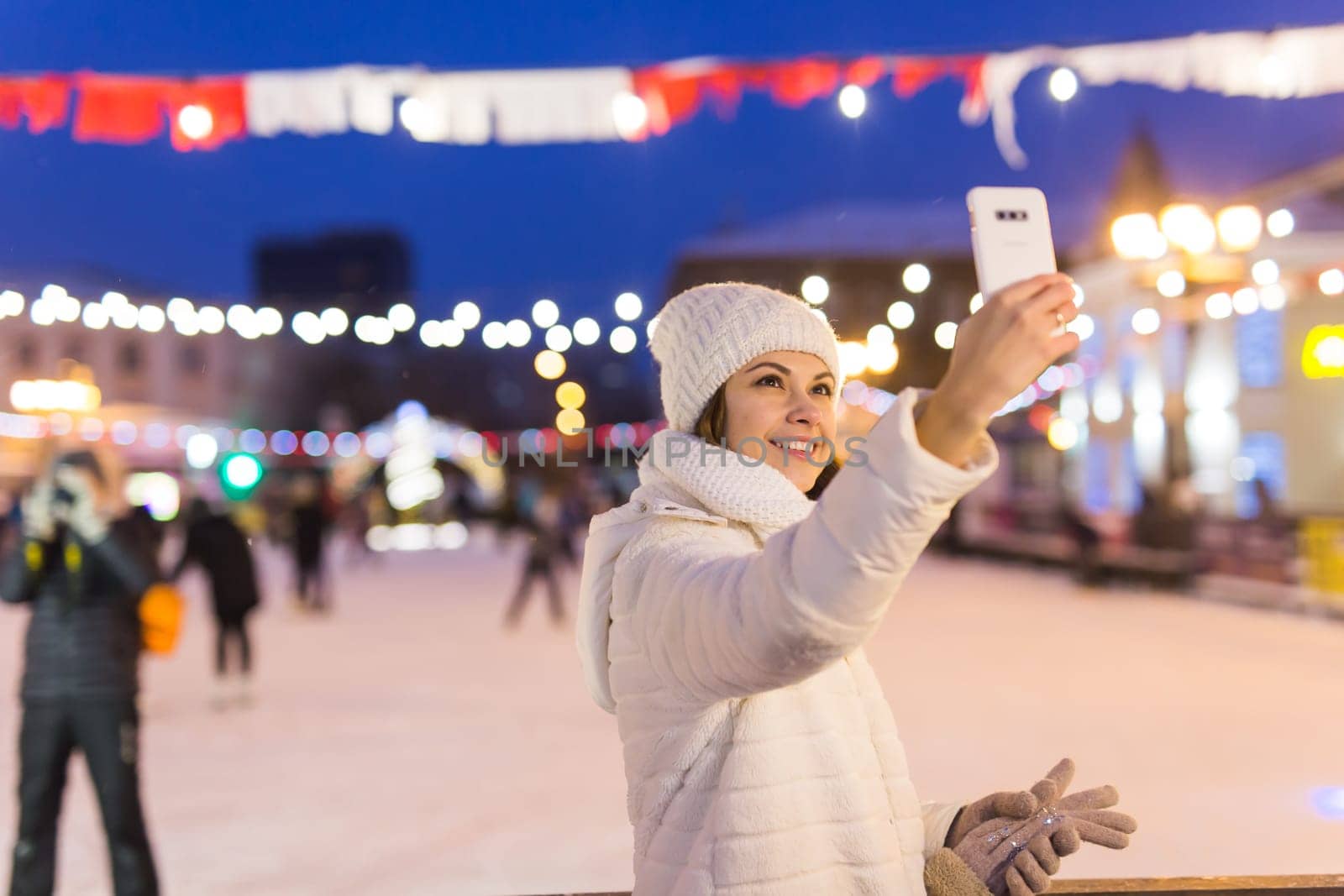 Christmas, winter, technology and leisure concept - happy young woman taking picture with smartphone on ice skating rink.