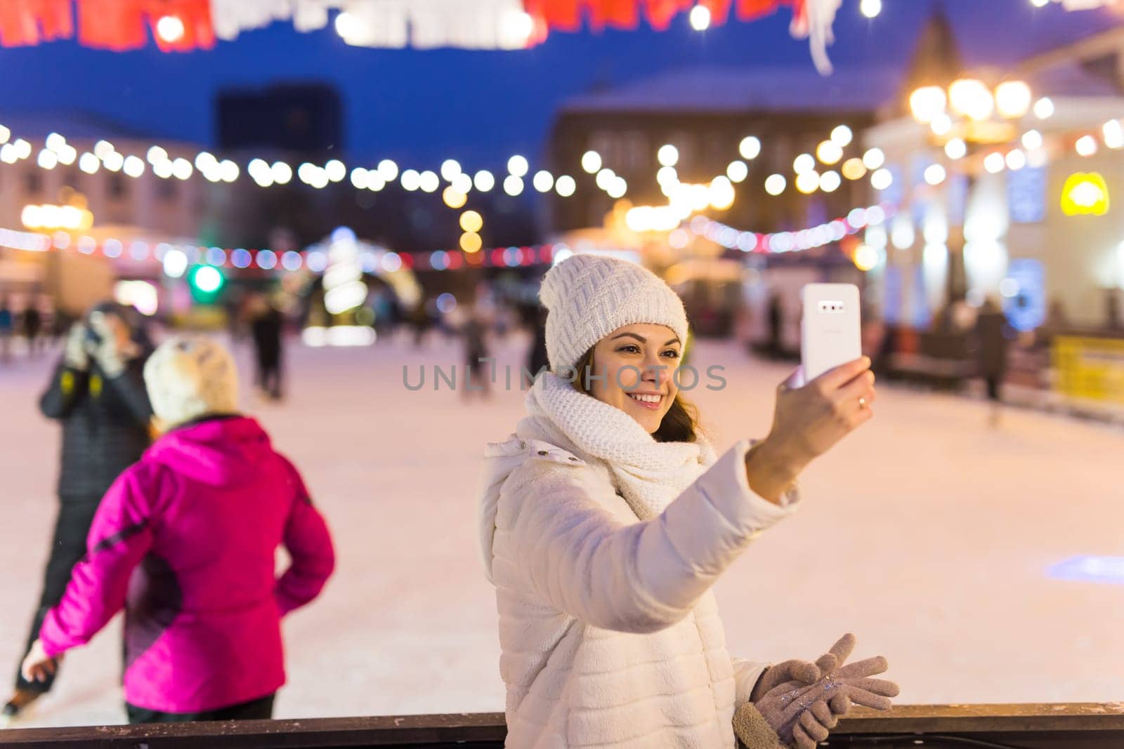 A woman on the rink is skating and taking selfie on smartphone. New Years Eve and Christmas. Fairy lights. Ice and snow mood concept. Winter
