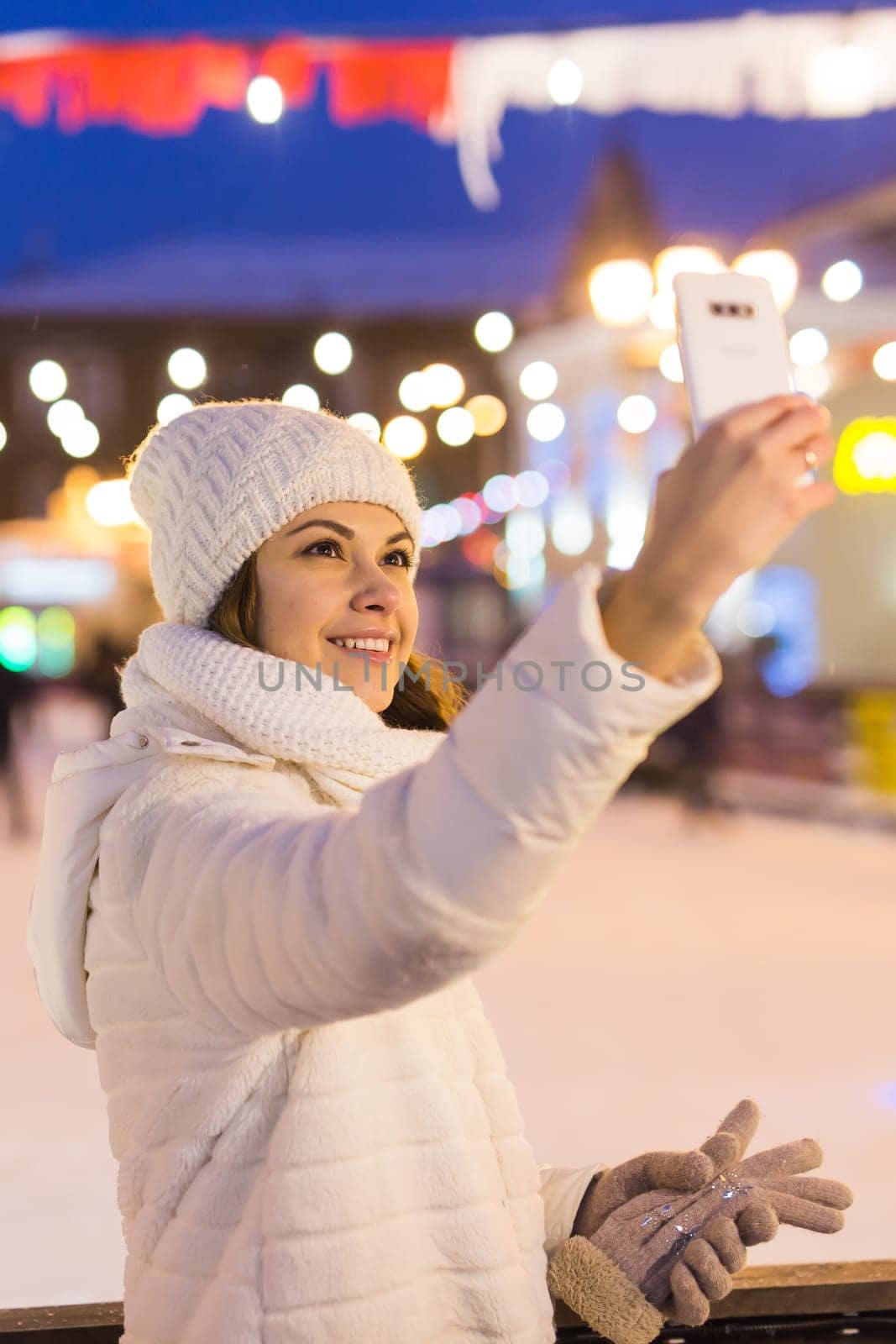 Christmas, winter, technology and leisure concept - happy young woman taking picture with smartphone on ice skating rink.