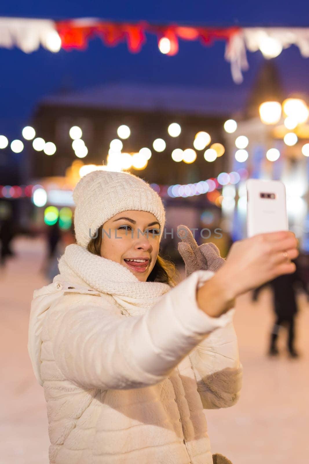 A woman on the rink is skating and taking selfie on smartphone. New Years Eve and Christmas. Fairy lights. Ice and snow mood concept. Winter